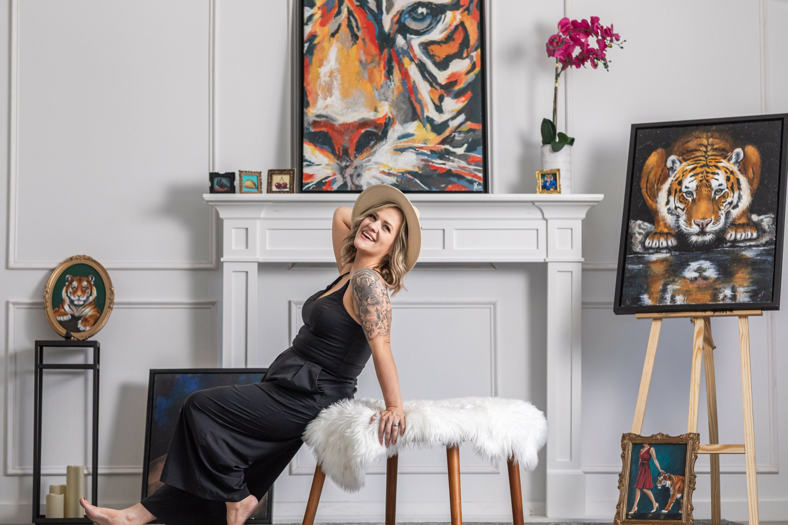 Woman in black dress sitting on a white furry bench in a decorated room with artwork and a fireplace.