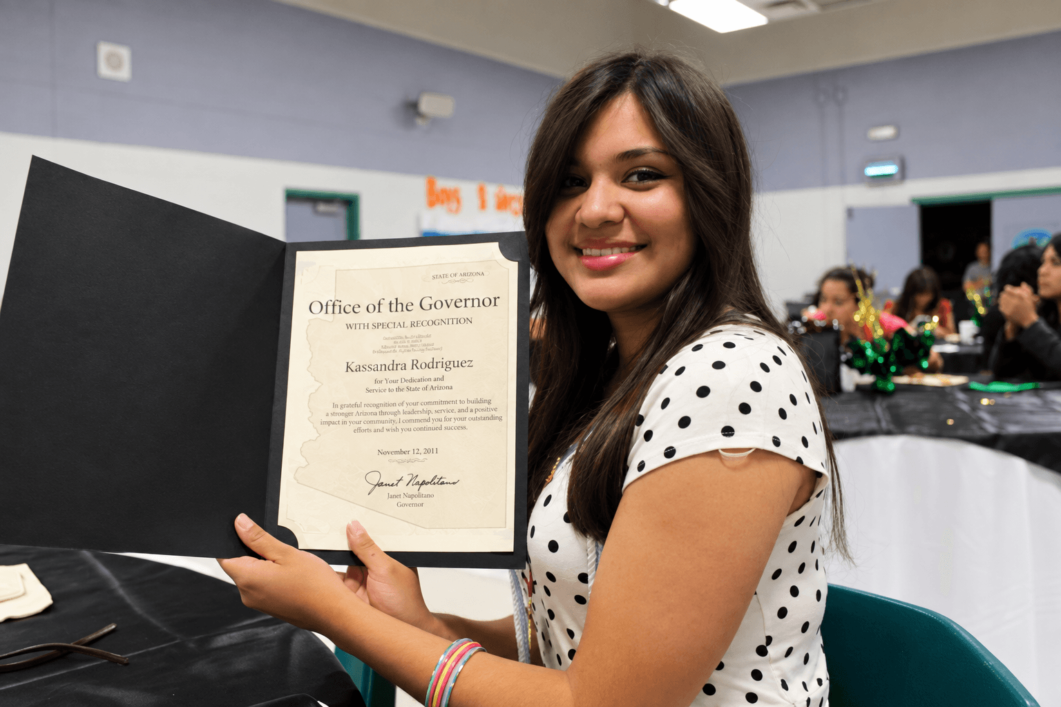 Young woman holding a certificate, smiling, in a room with people and tables in the background.
