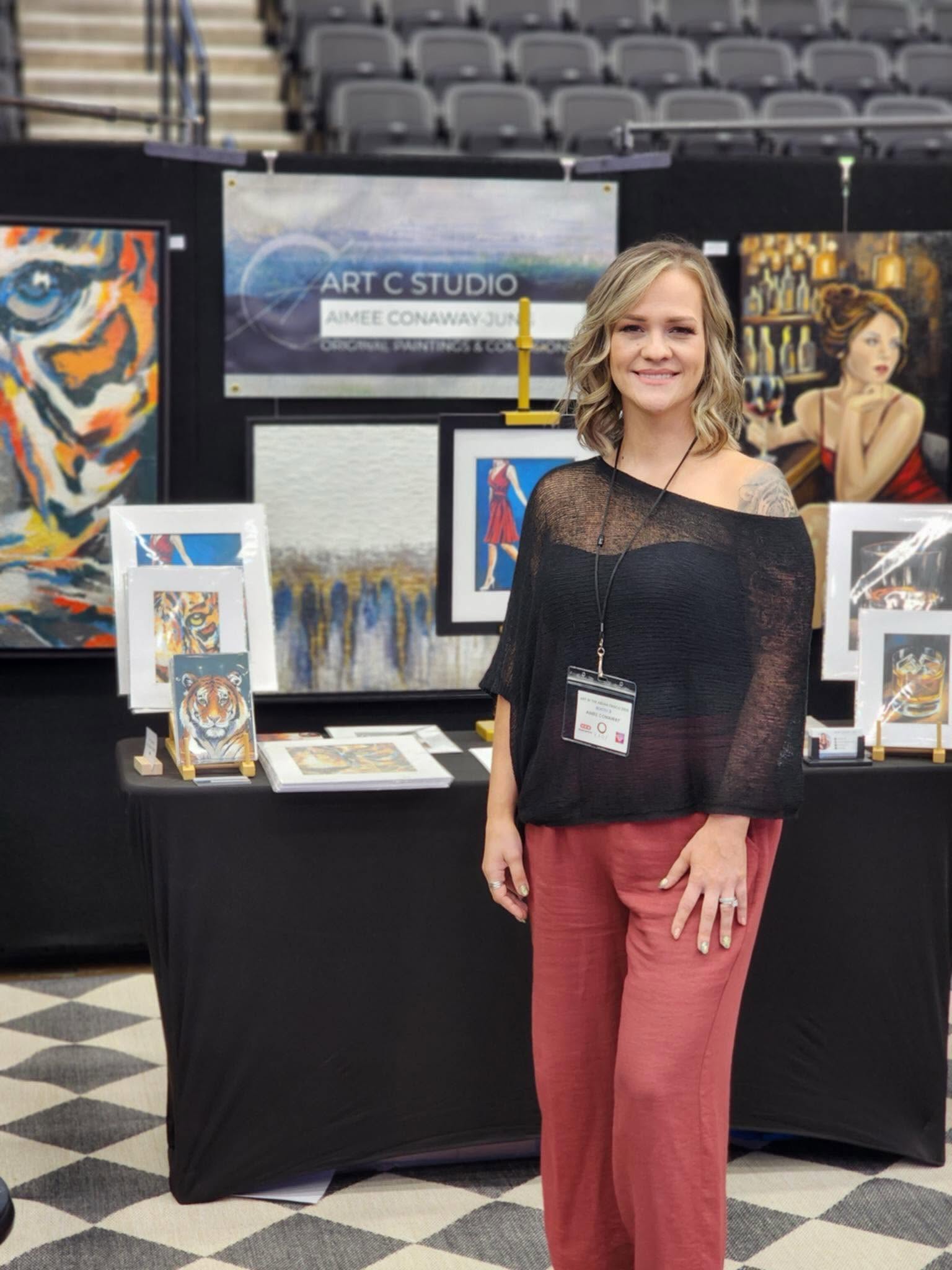 Woman with blonde hair smiling at art display booth, wearing black top and red pants, in an indoor exhibition space.