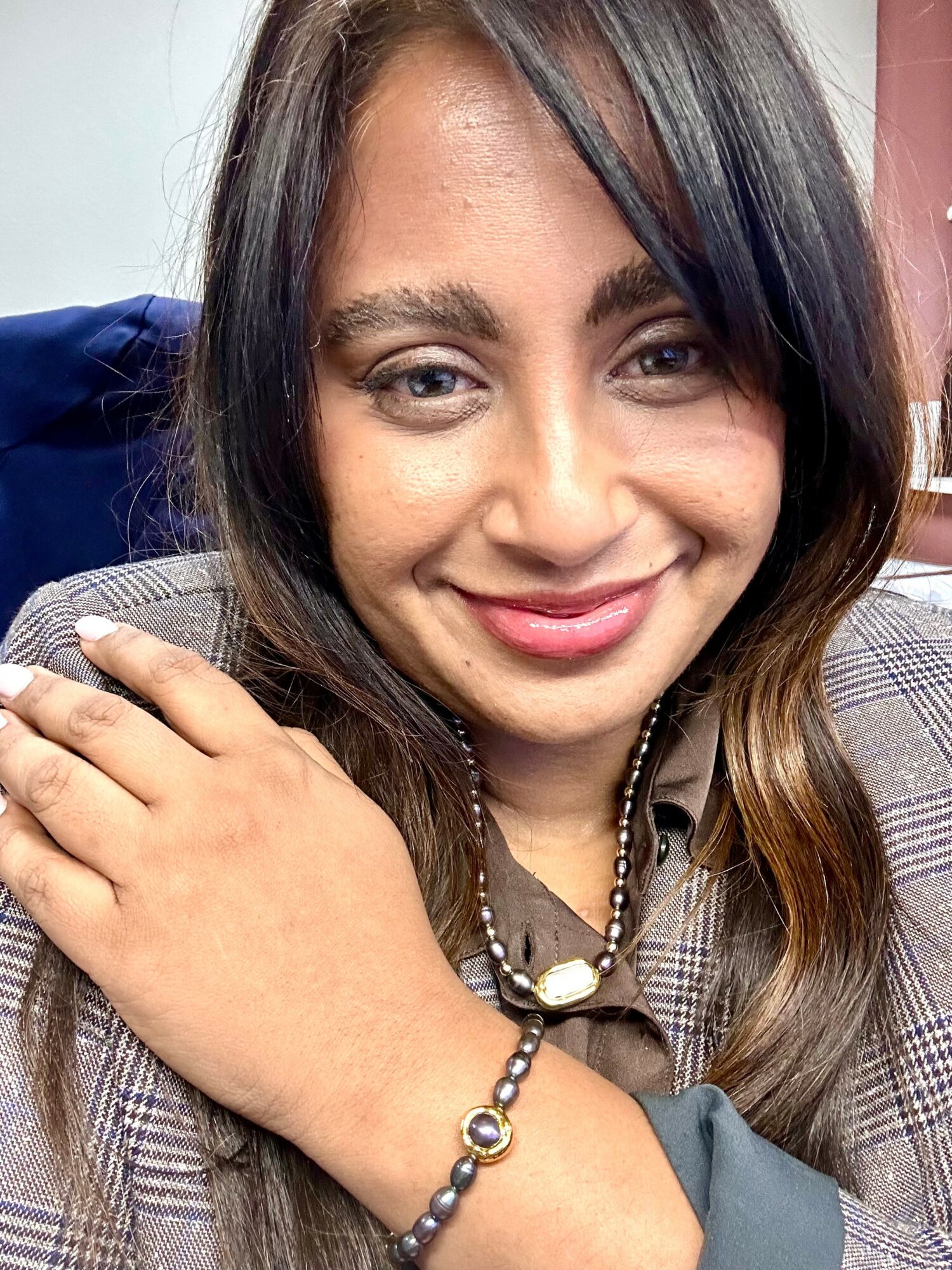 Close-up of a smiling woman with long dark hair, wearing a plaid blazer and jewelry, resting her hand on her shoulder.