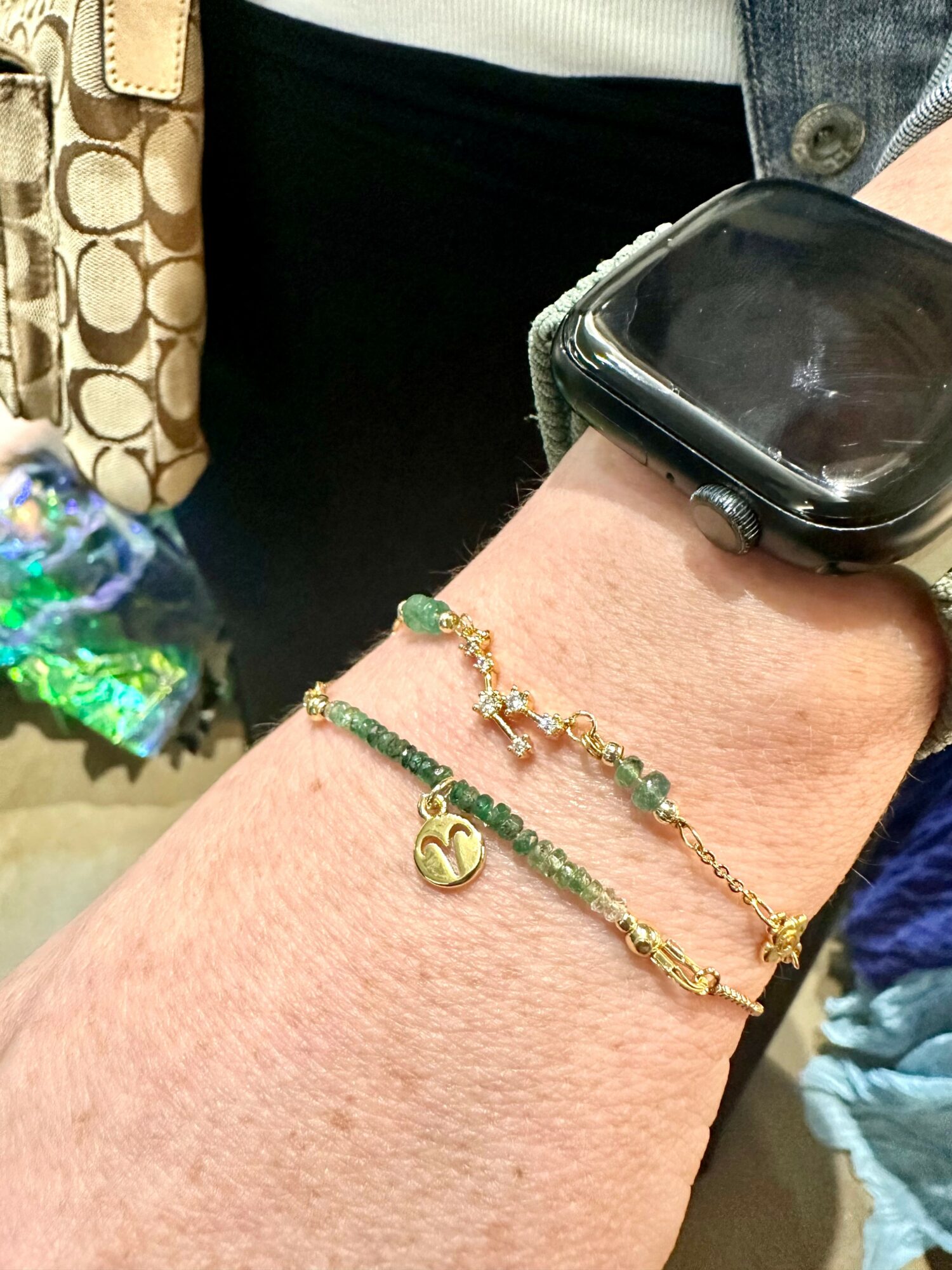Person's wrist with a smartwatch and two bracelets, one gold with a heart charm, and one green with beads.