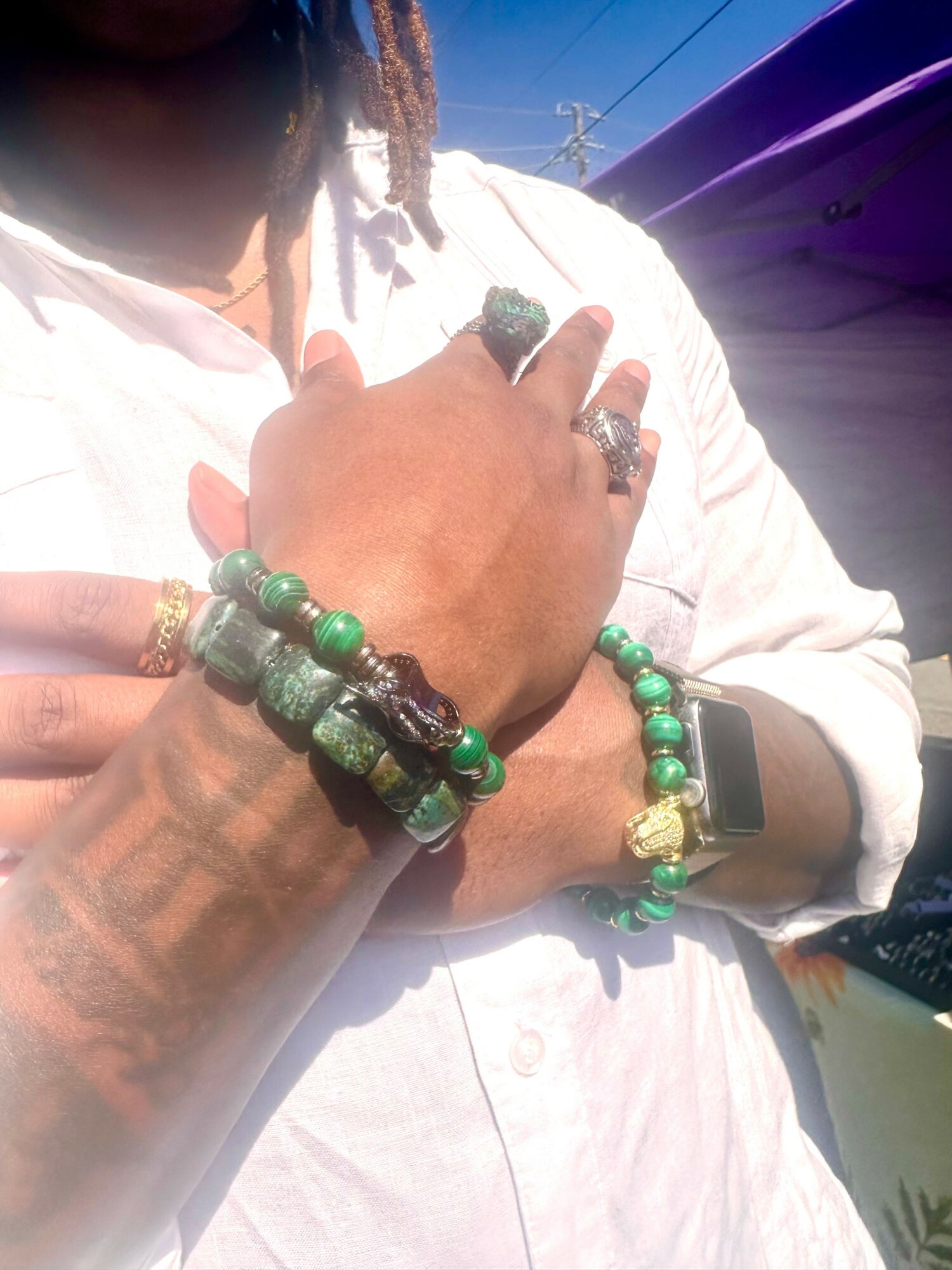 Person wearing multiple green and black beaded bracelets, rings, and a watch, with a white shirt and outdoor background.