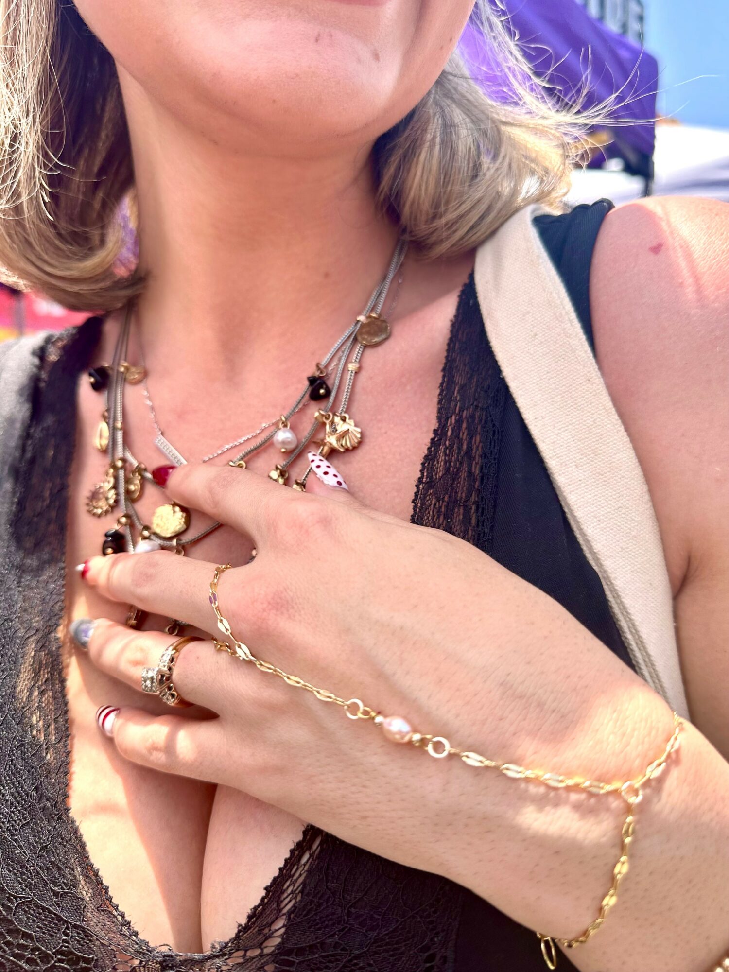 Close-up of a woman wearing layered necklaces and rings, with her hand resting on her chest, in a black top and beige vest.