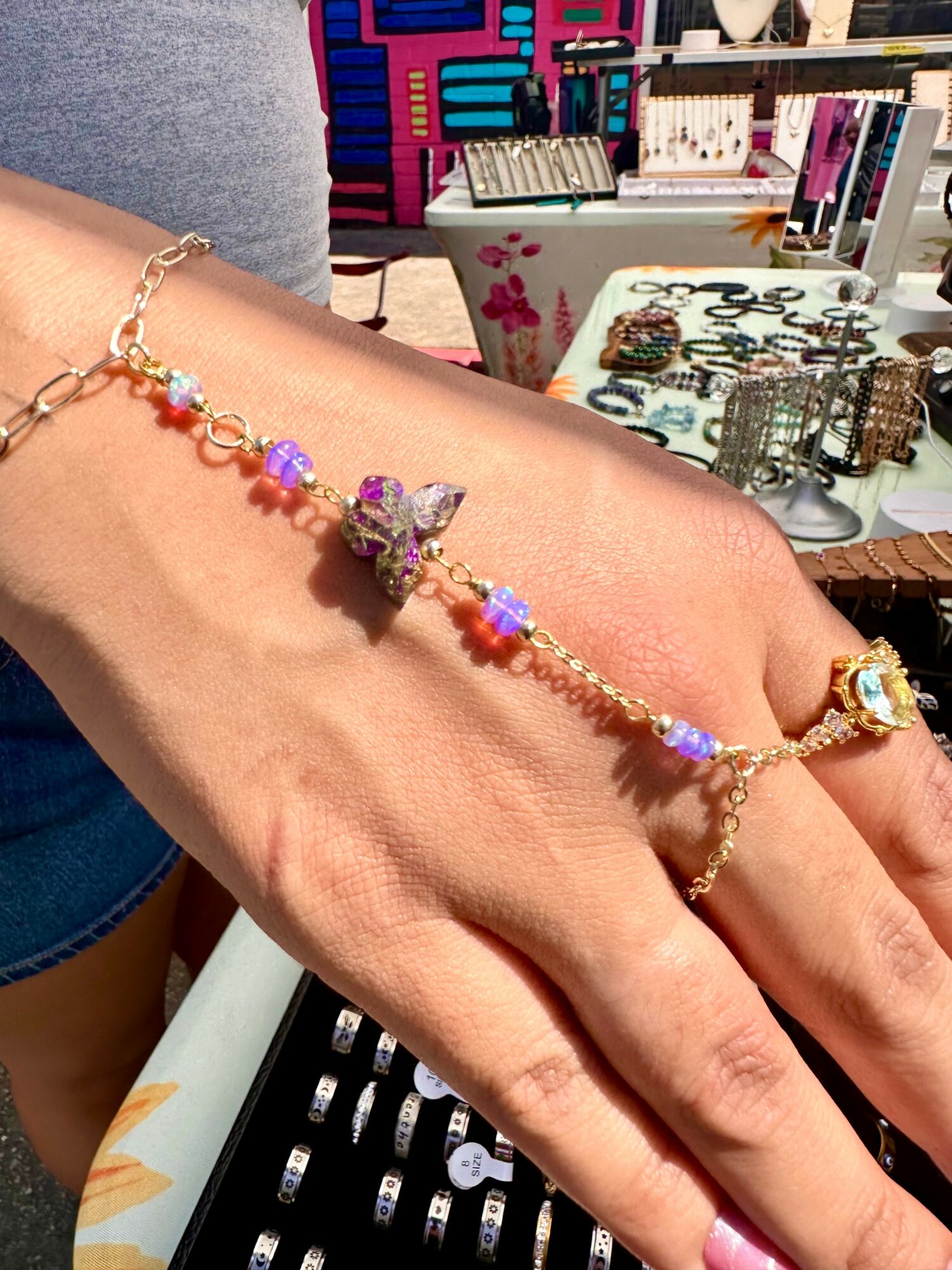 Person's hand with a colorful beaded bracelet, jewelry display table in background, outdoor market setting.