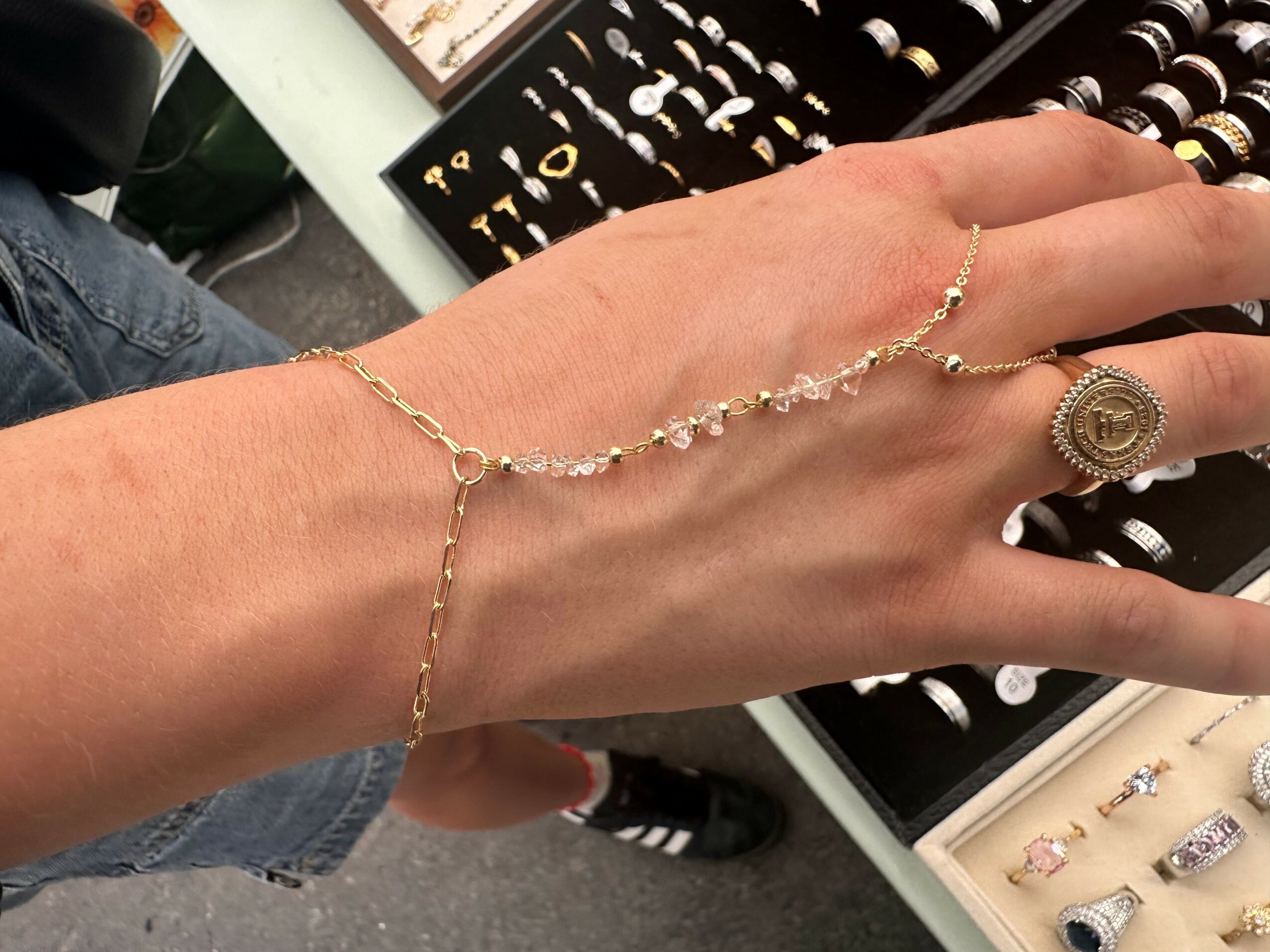 A hand with a gold bracelet and a large ring, displayed against a jewelry store background.
