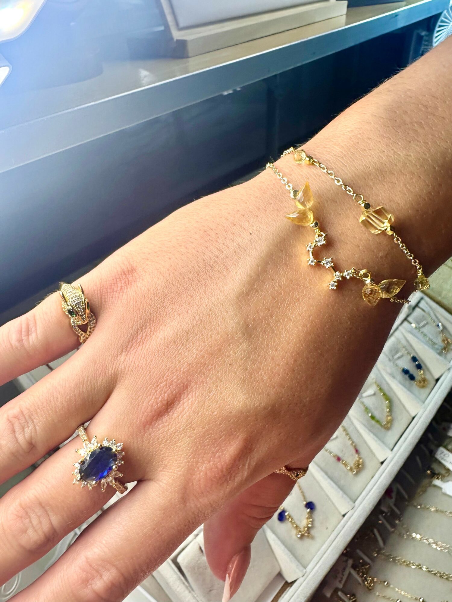 Hand with multiple gold rings and bracelets, jewelry featuring blue and clear stones, in a jewelry store display case.