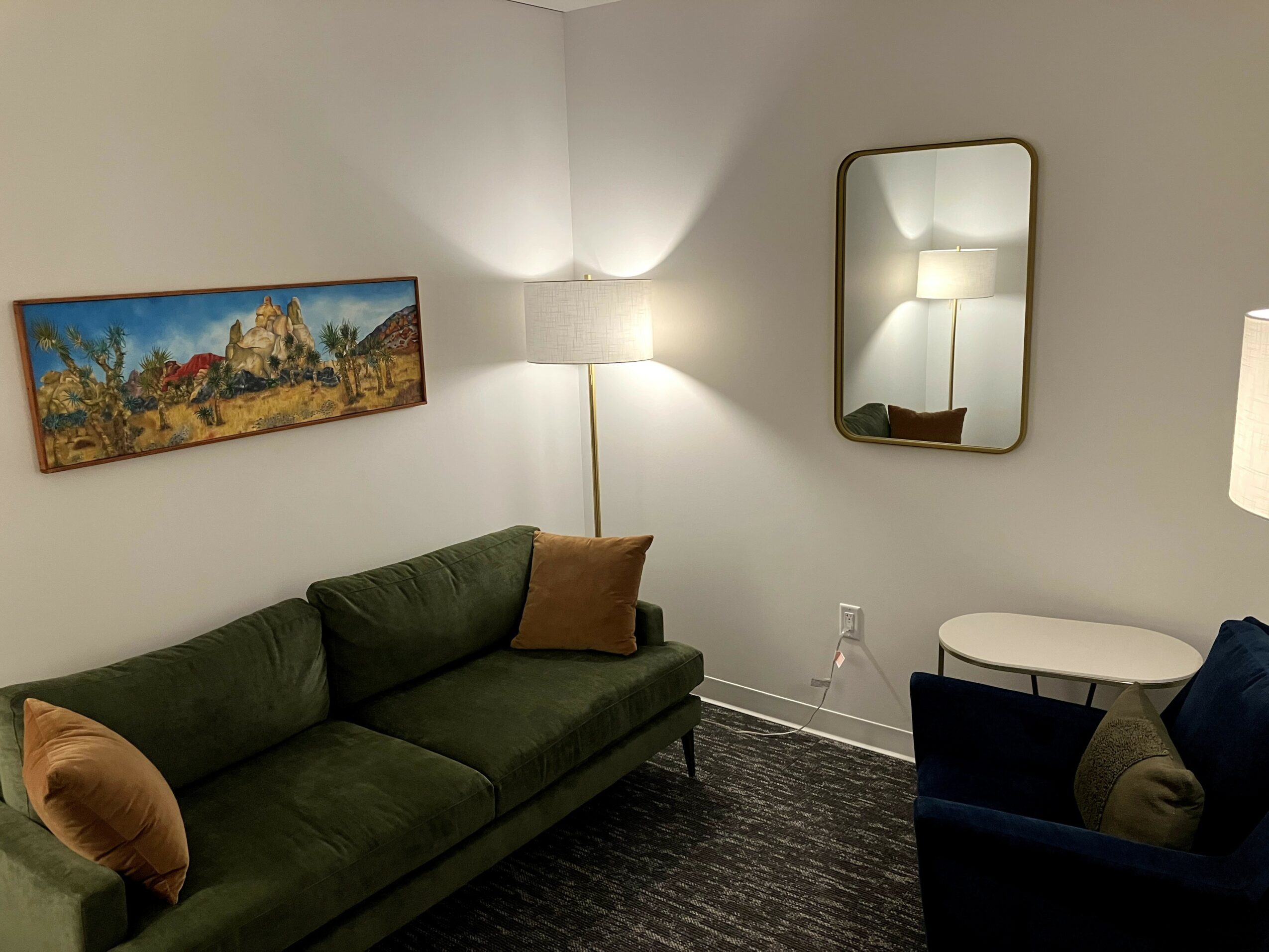 Living room with a green sofa, a brown pillow, a white lamp, a mirror, and a painting on the wall.