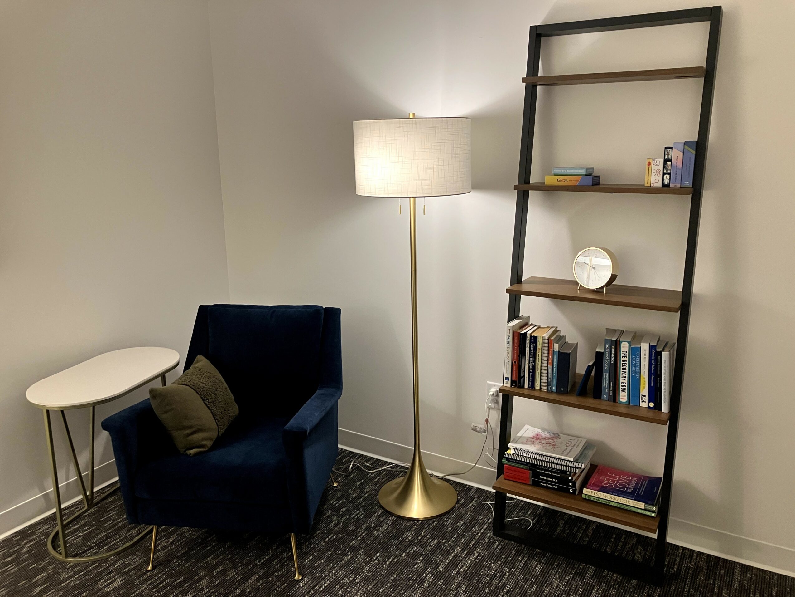 Corner of a room with a blue armchair, a small white side table, a tall floor lamp, and a black ladder-style bookshelf with books and decor. Word count: 27