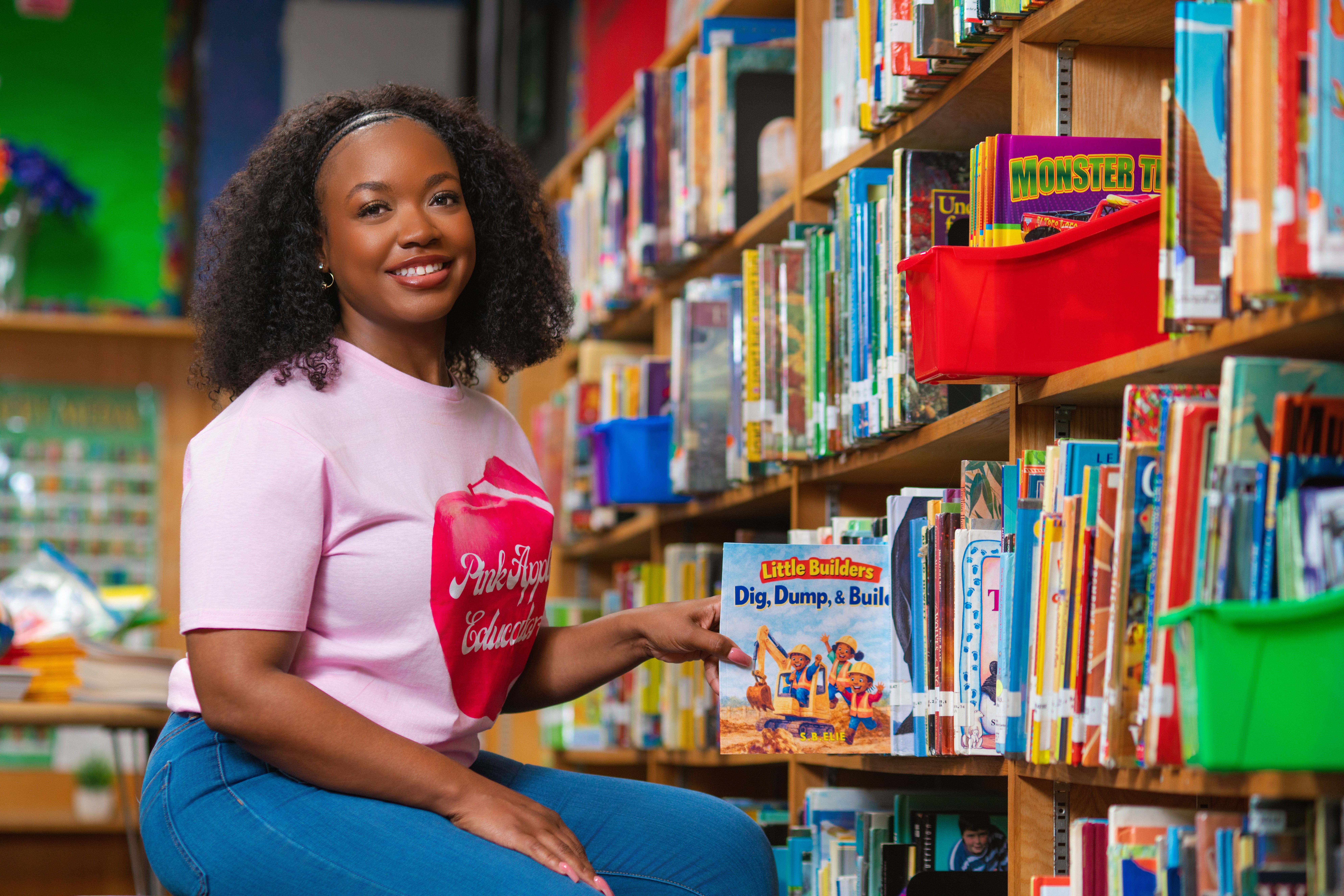 Young girl with curly hair smiling in a library, holding a book, surrounded by colorful books on shelves.