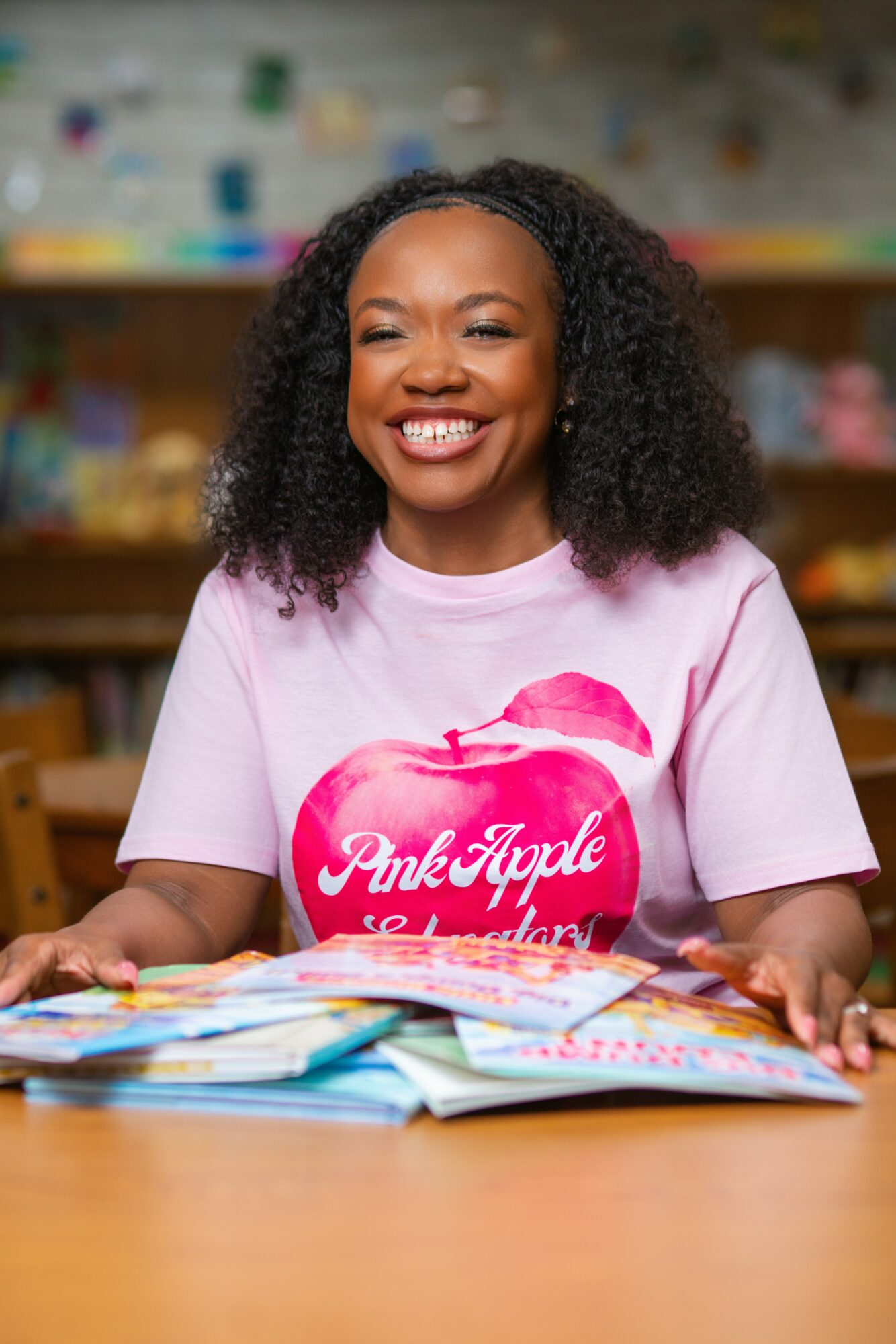 Smiling woman with curly hair wearing a pink T-shirt with a pink apple graphic, sitting at a table with colorful papers.