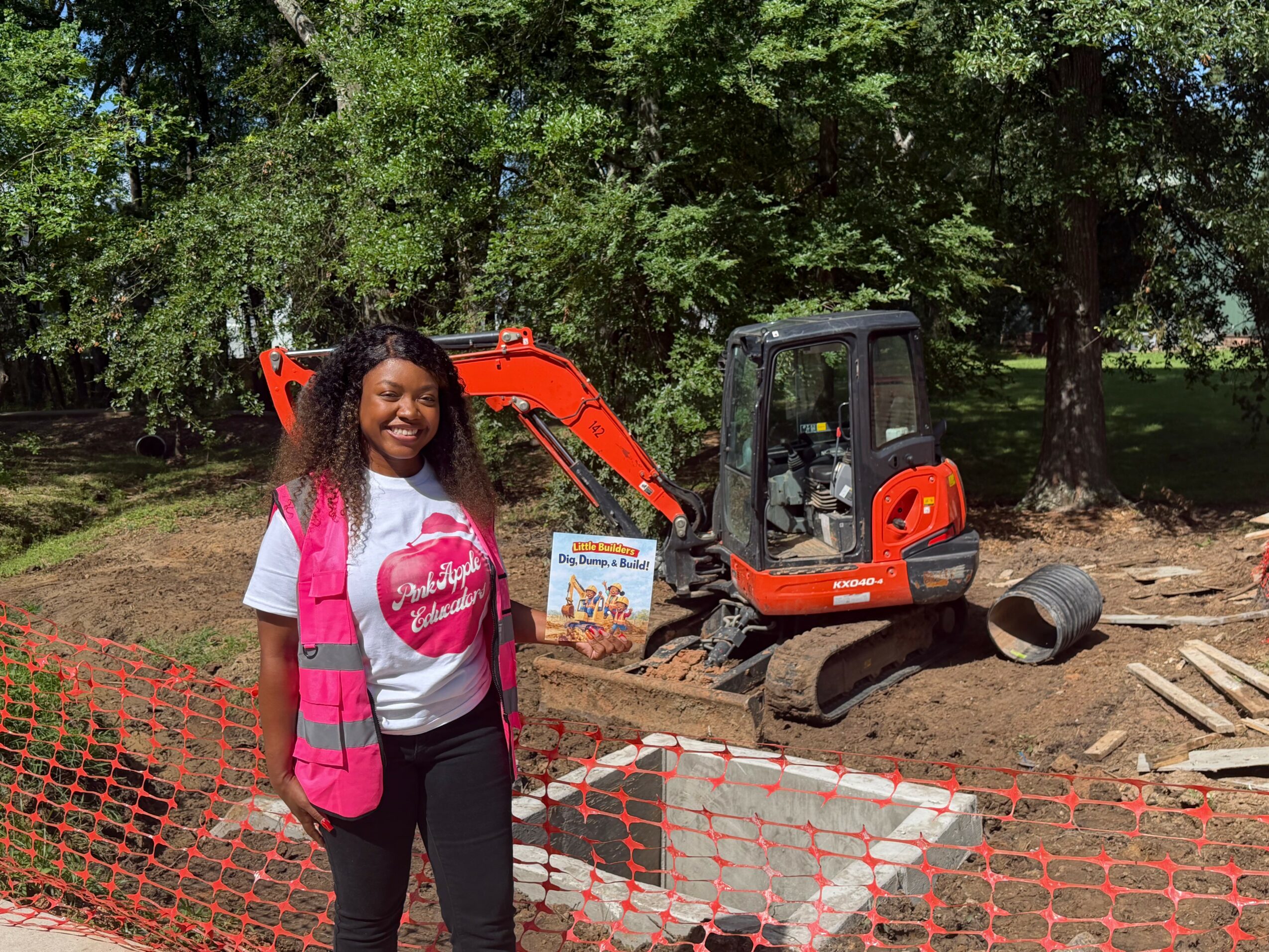 Smiling woman in pink safety vest and white shirt standing outdoors near construction site with small excavator and trees.