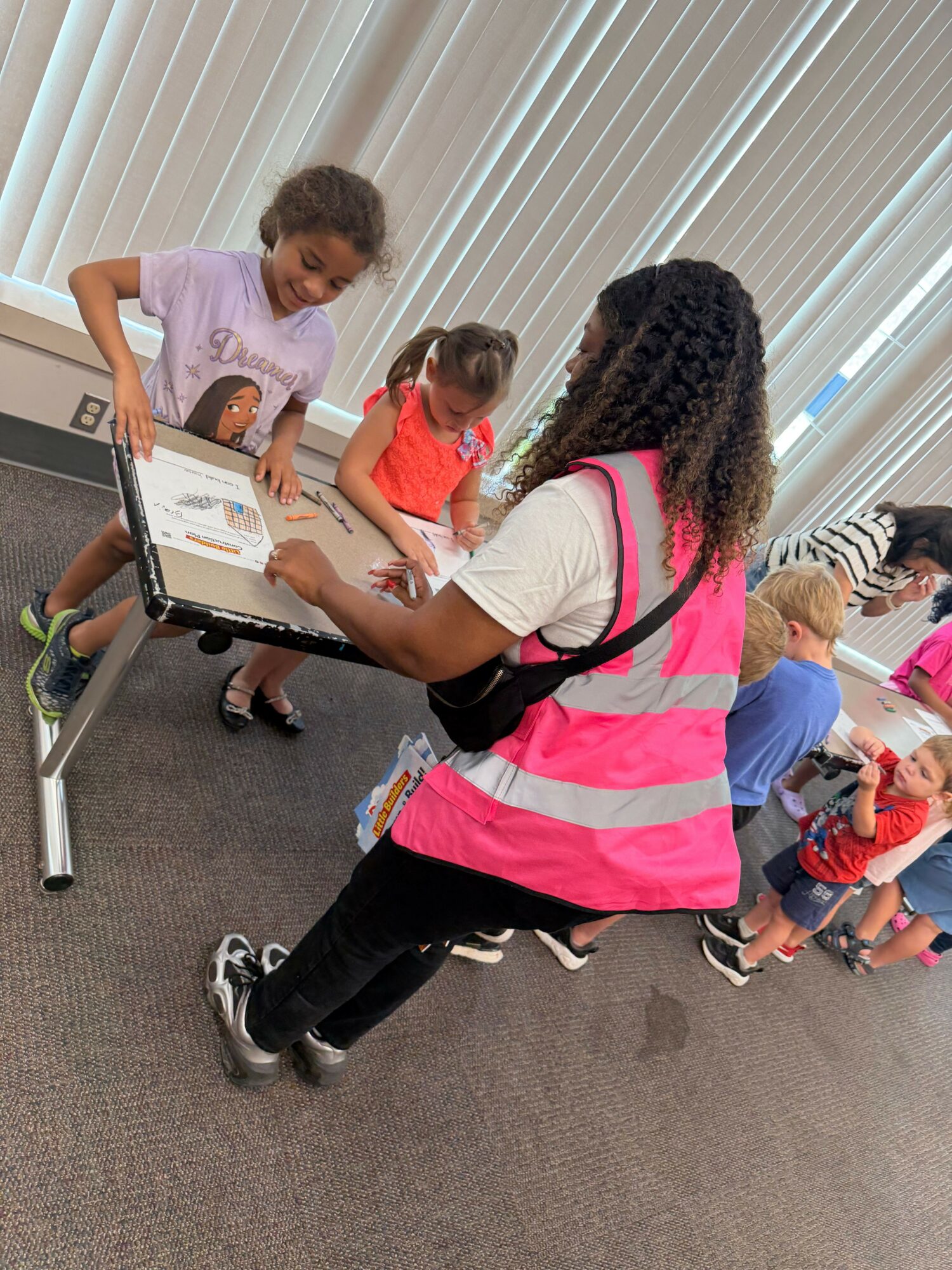 Adult woman in a pink safety vest assisting a young girl with a laptop, children in background, indoor setting.