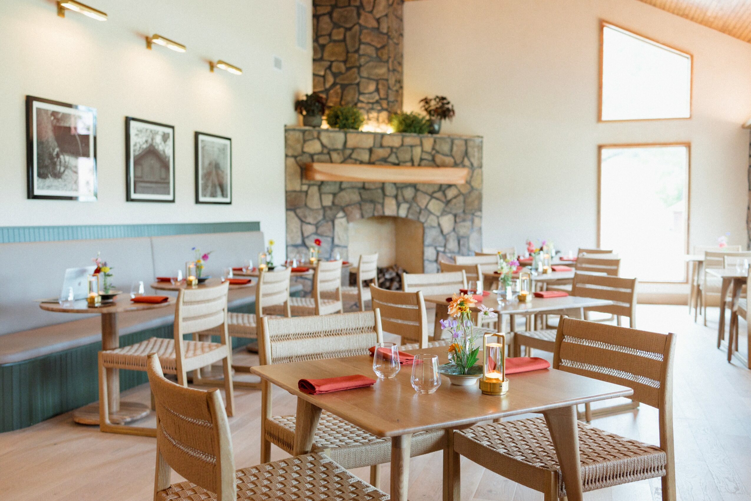 Dining area with wooden tables, chairs, a stone fireplace, and large windows, decorated with flowers and framed pictures on the wall.