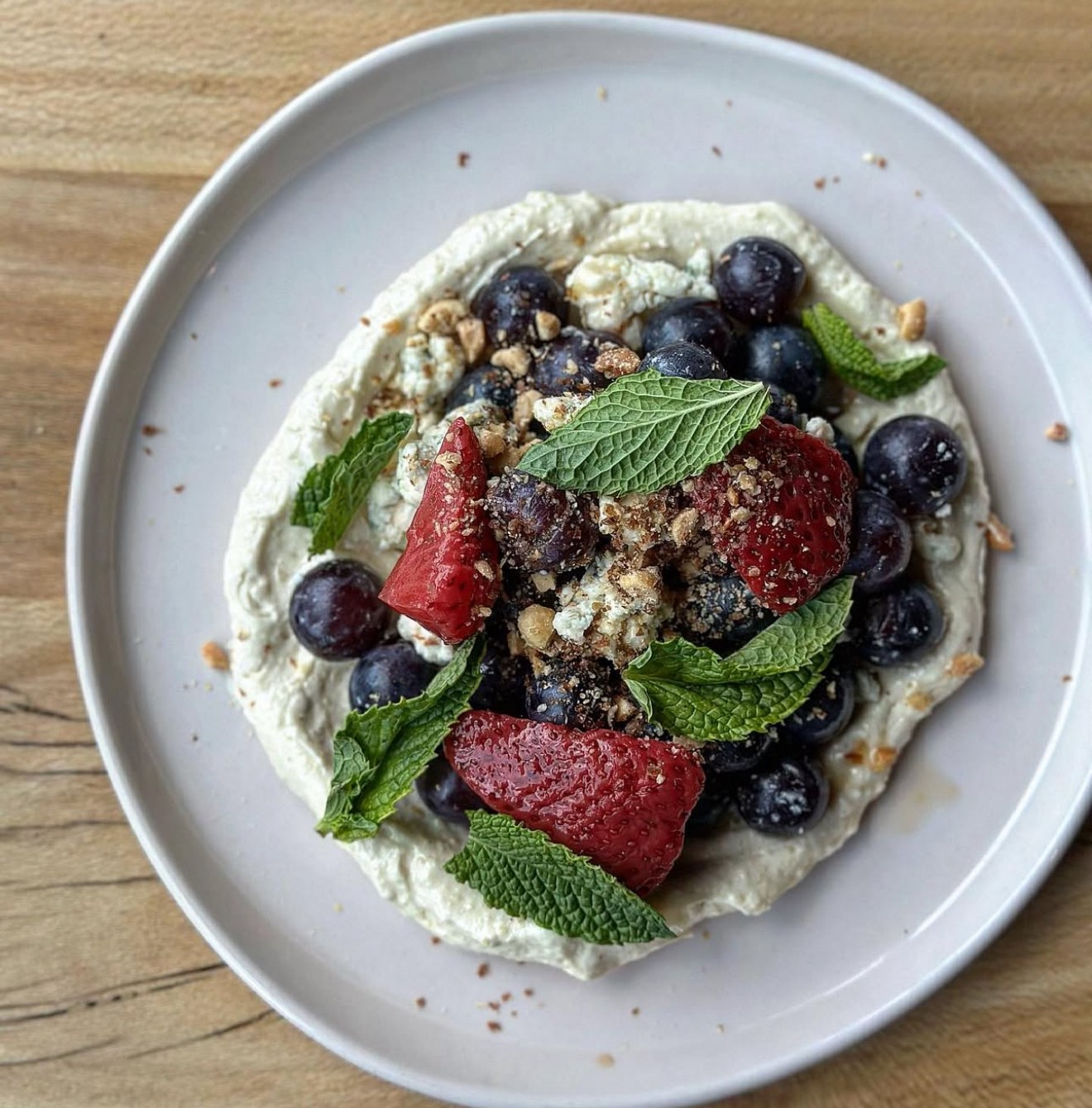 Plate with yogurt, blueberries, strawberries, mint leaves, and nuts on a wooden surface.