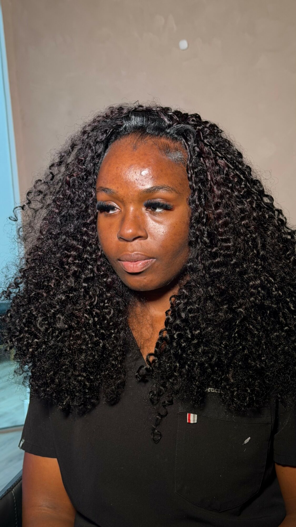 Woman with curly hair looking to the side, wearing a black shirt, indoors with beige wall background.