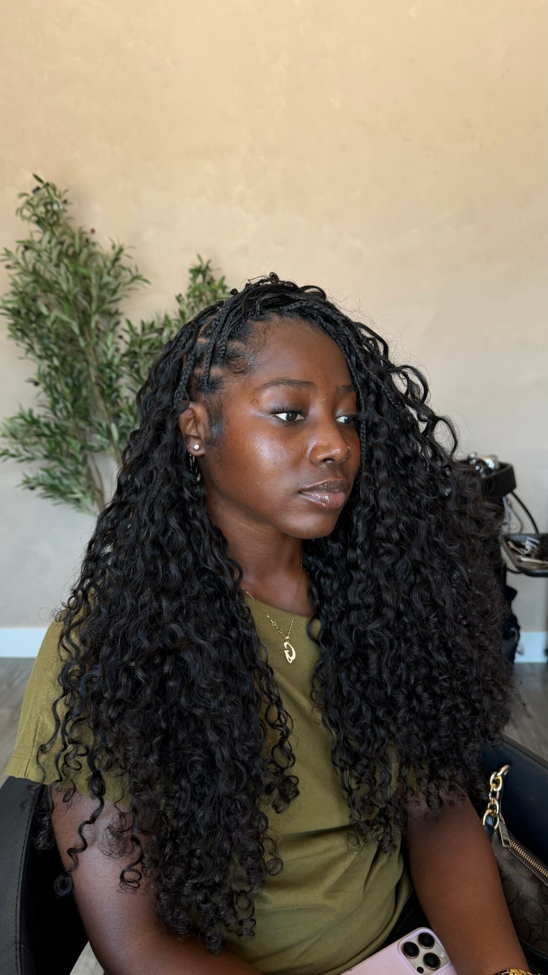 Young woman with long curly hair wearing an olive green top, sitting indoors with a plant in the background.