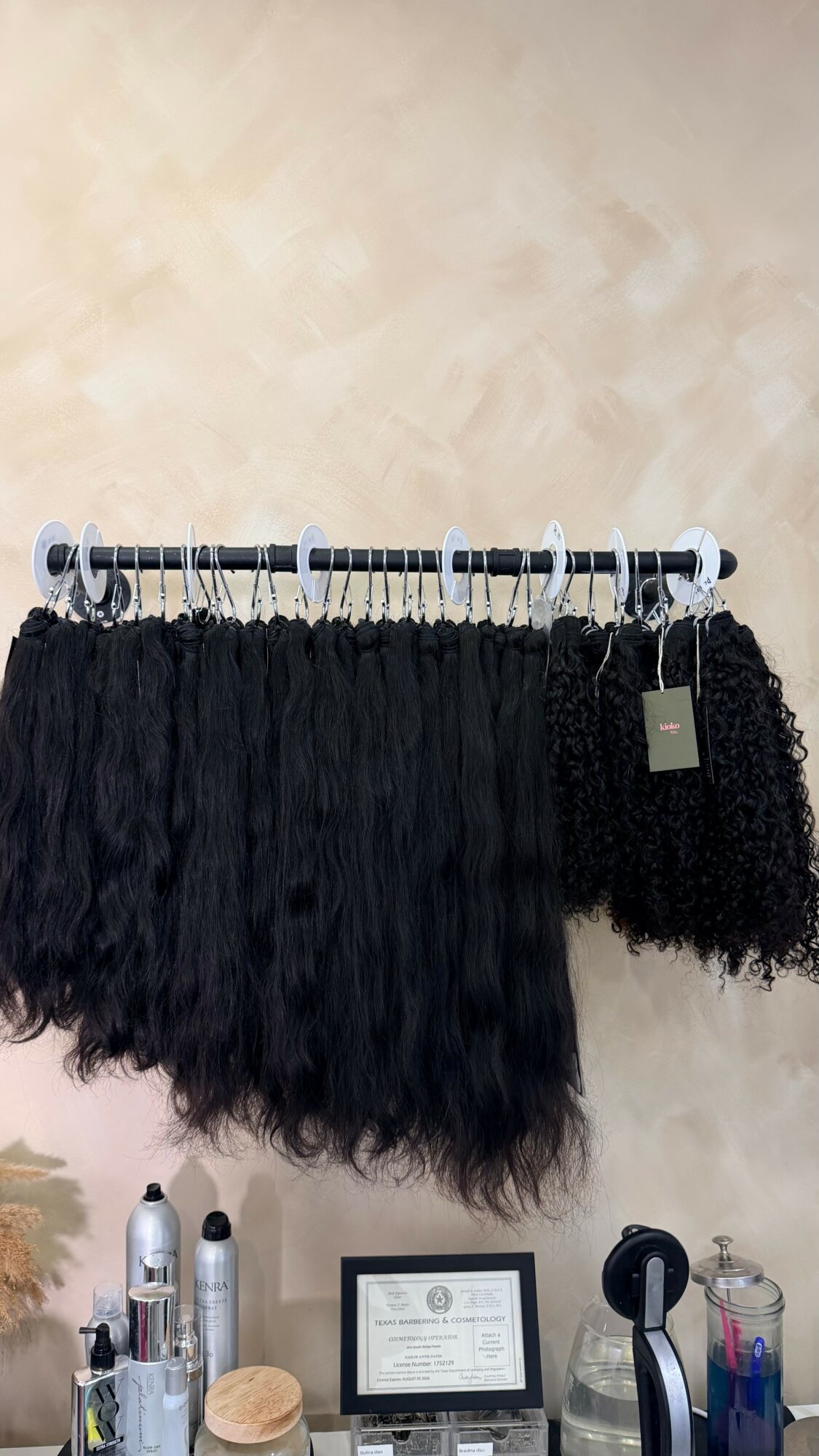 Black curly hair extensions hanging on a white rod, with a tag attached, in a room with a beige wall.