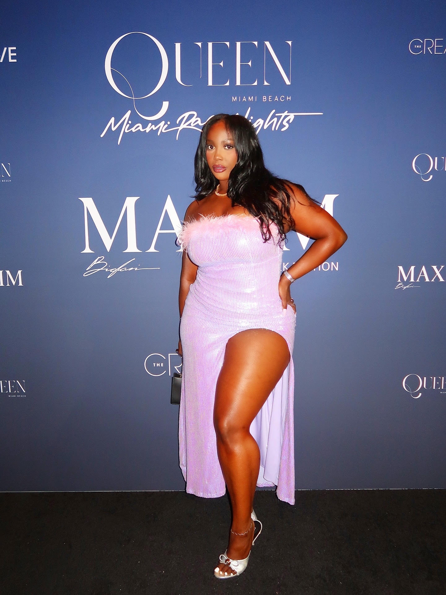 Woman in a pink dress with a high slit, standing in front of a blue backdrop with text, posing with one hand on her hip.