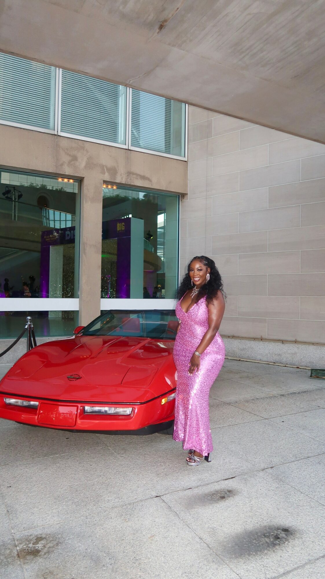 Woman in pink dress standing next to a red sports car outside modern building, large windows, concrete walls.