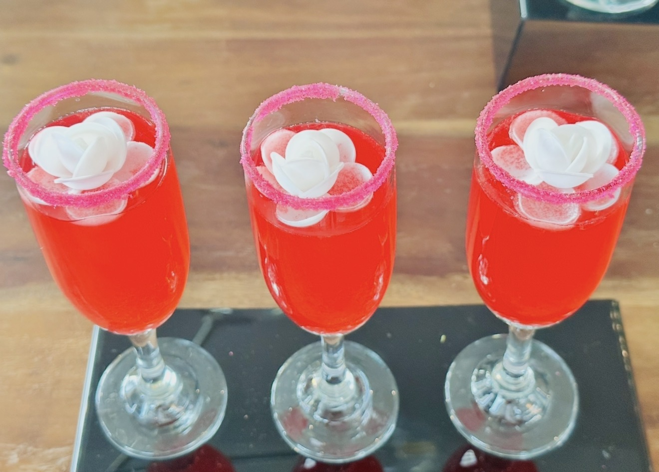 Three red drinks in tall glasses with white flower garnishes on a black tray, on a wooden surface.