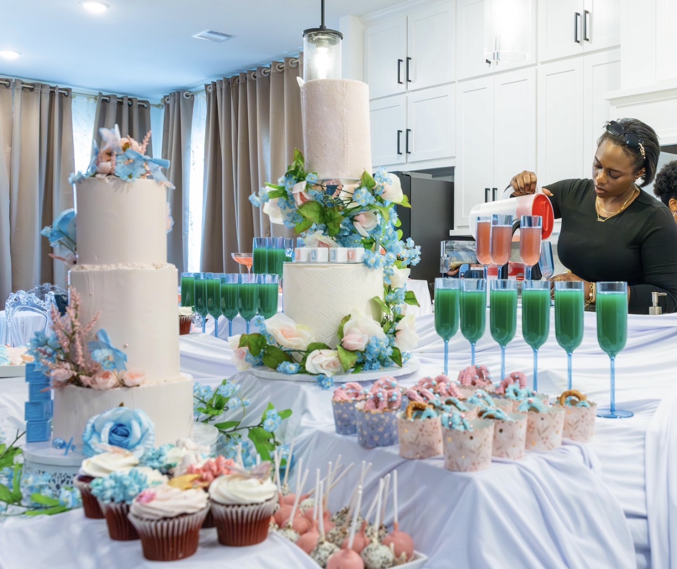 Woman pouring pink drink at decorated table with cakes, cupcakes, and floral arrangements, in a bright room with curtains.