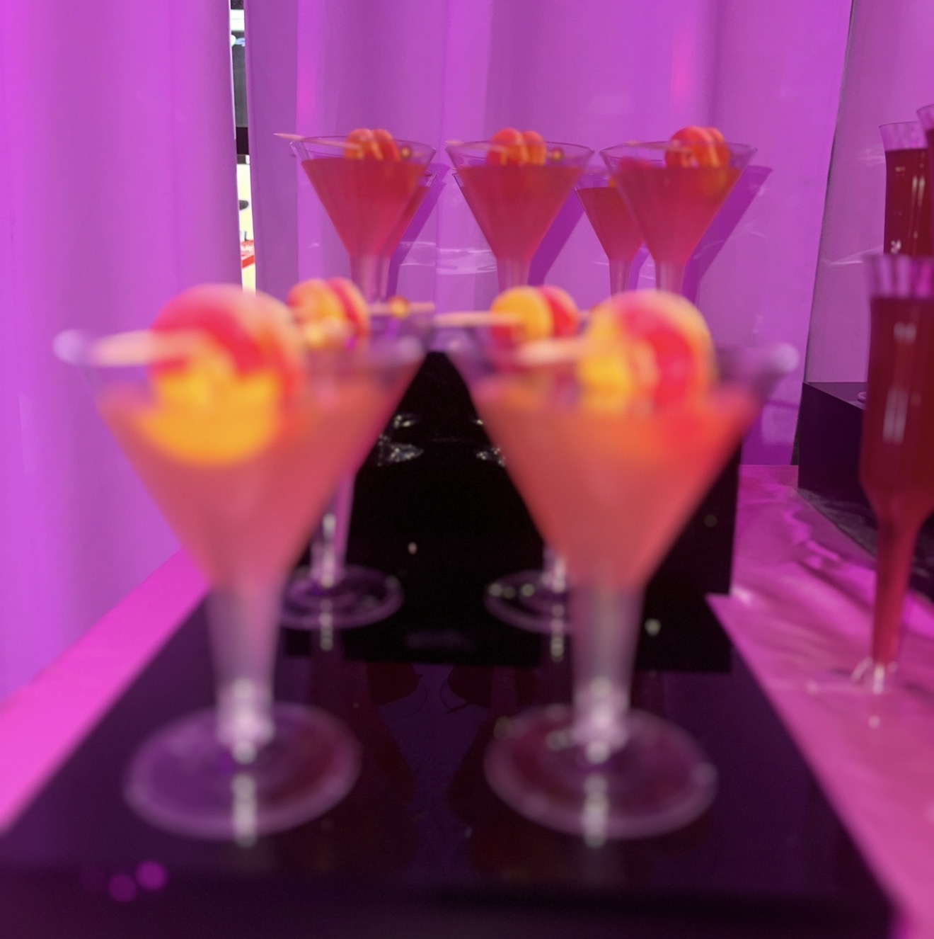 Six pink cocktails with skewered fruit garnishes on a black tray, pink lighting in the background.
