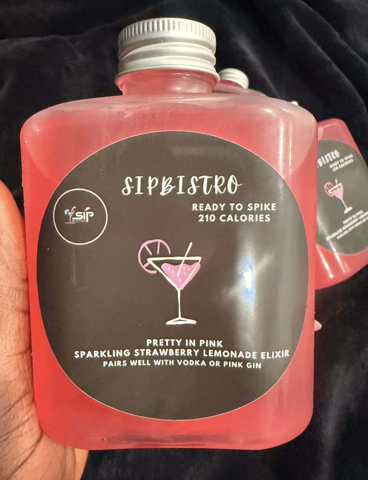 Pink bottle with a black label featuring a cocktail glass illustration and text about a strawberry lemonade elixir.