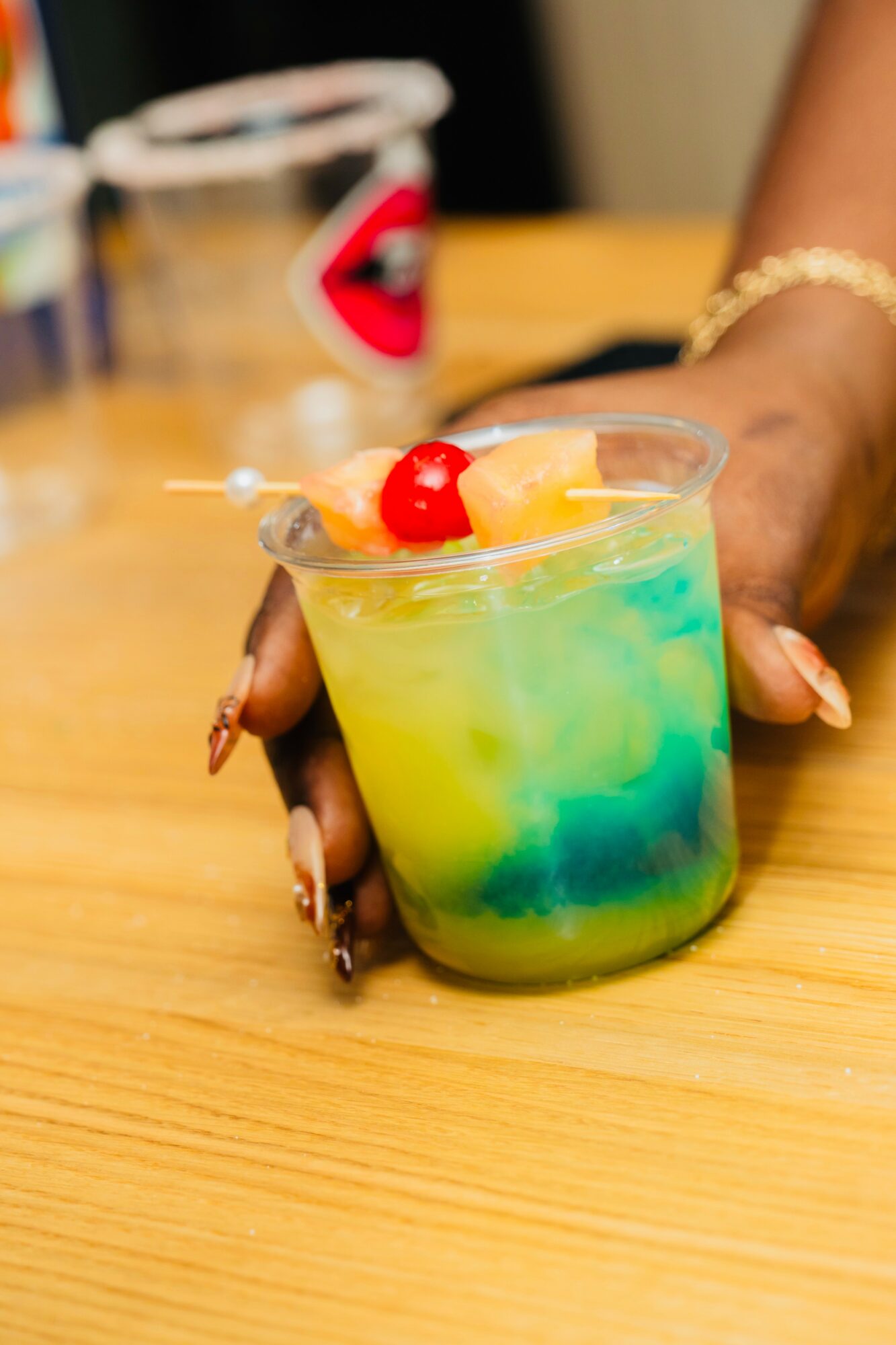 Hand holding a colorful layered cocktail with cherries and lemon slice on a wooden table.