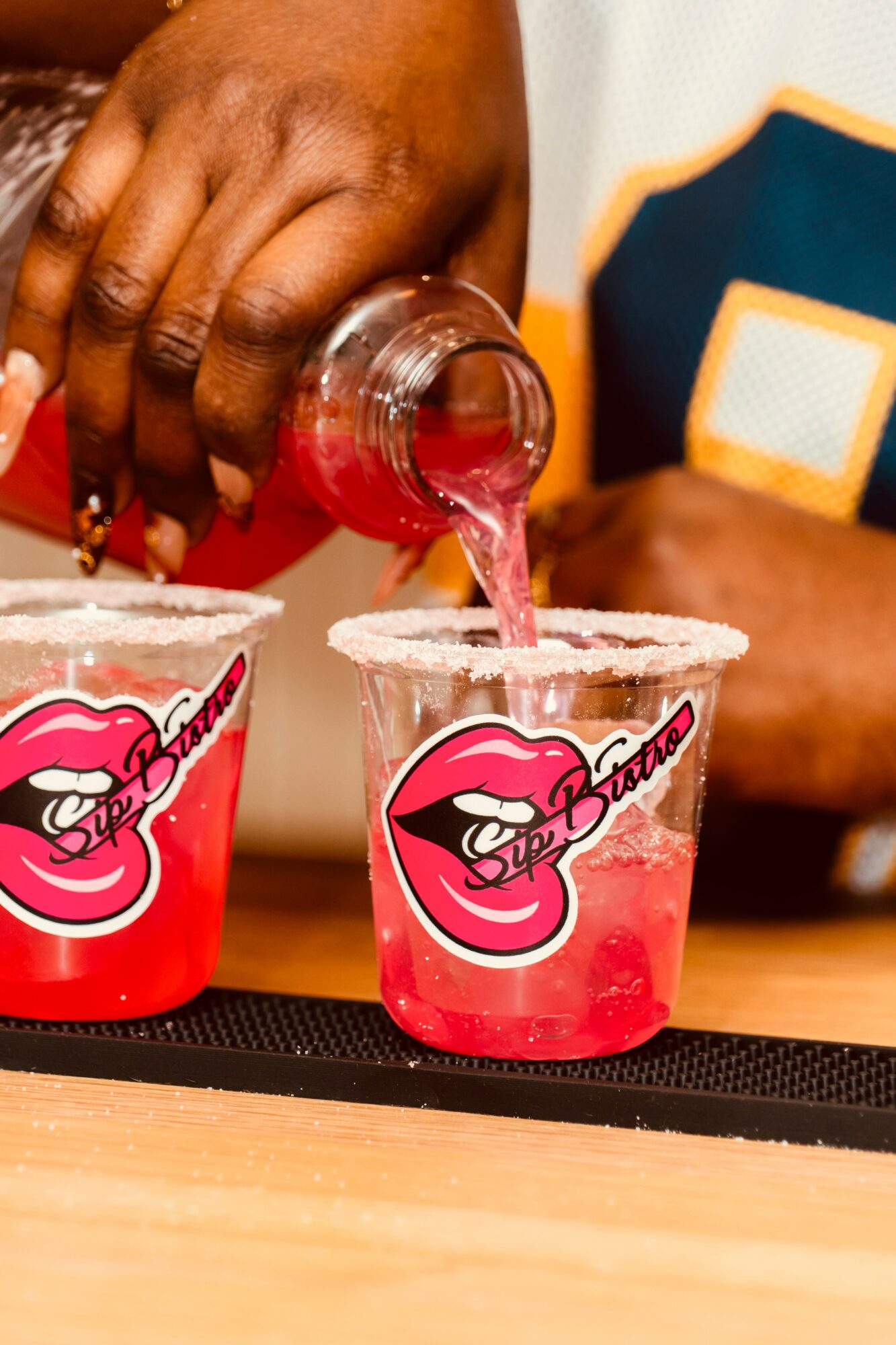 Person pouring pink drink into cups with lips logo, salt rim, and a hand in the background, close-up view.