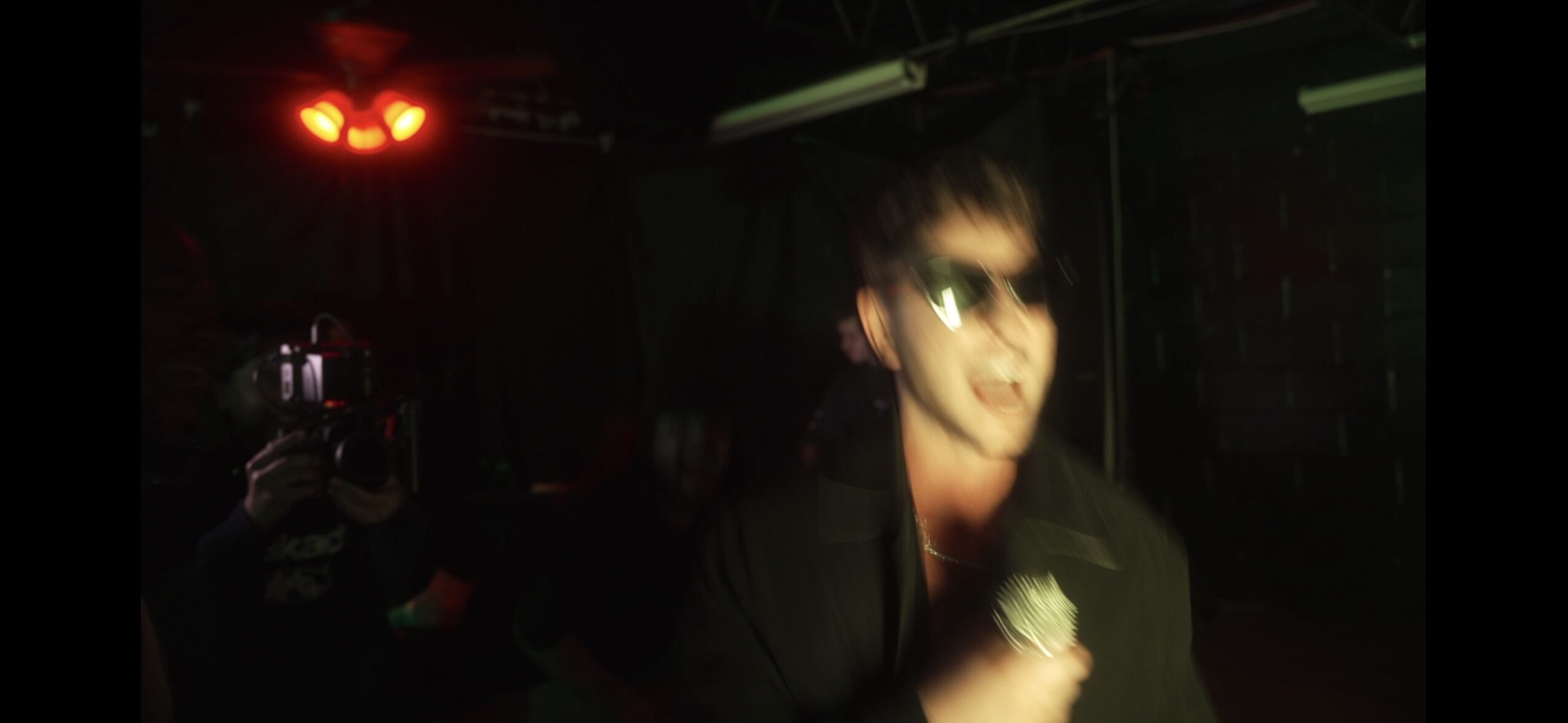 Person with sunglasses singing or speaking into a microphone in a dark setting.