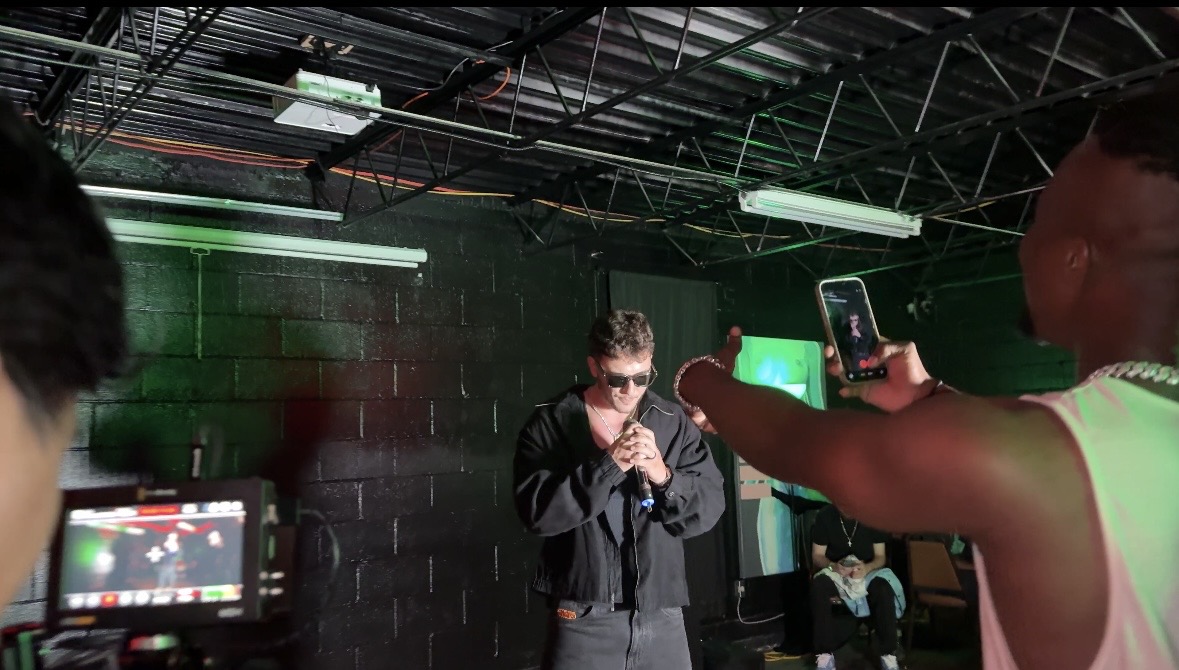 Person in dark clothing stands in front of a black wall with green lighting, holding a microphone, others record with phones.