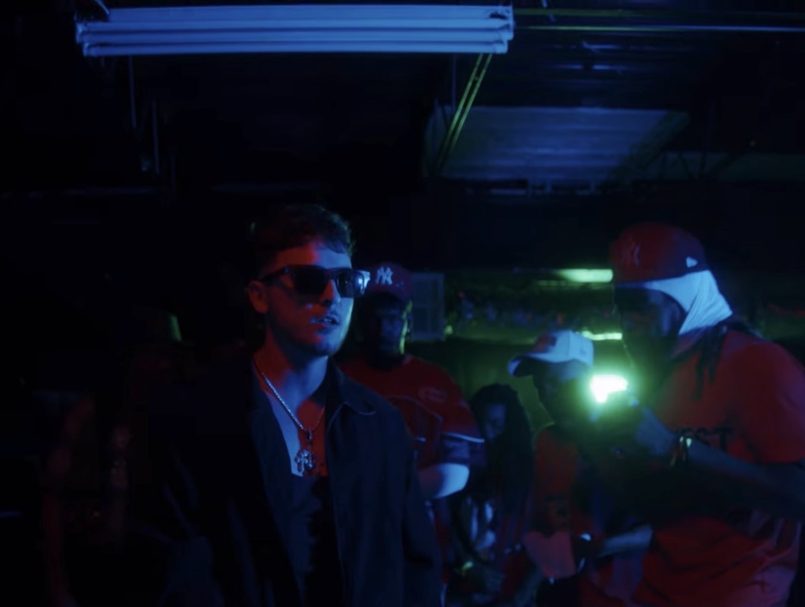 People in a dark room with neon lighting, one person wearing sunglasses and another with a cap, engaging in conversation.