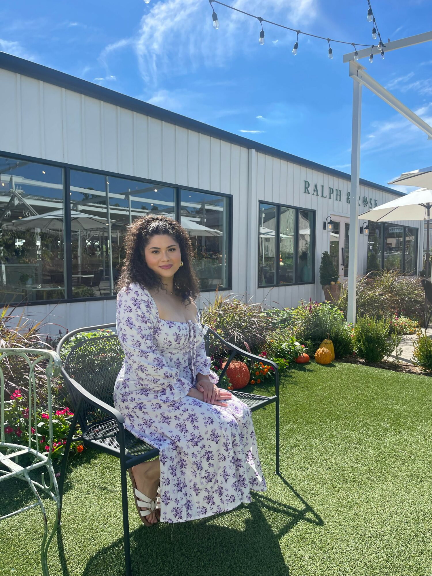Young woman with curly hair in a long floral dress sitting on a black metal bench outdoors, with a building and garden in background.
