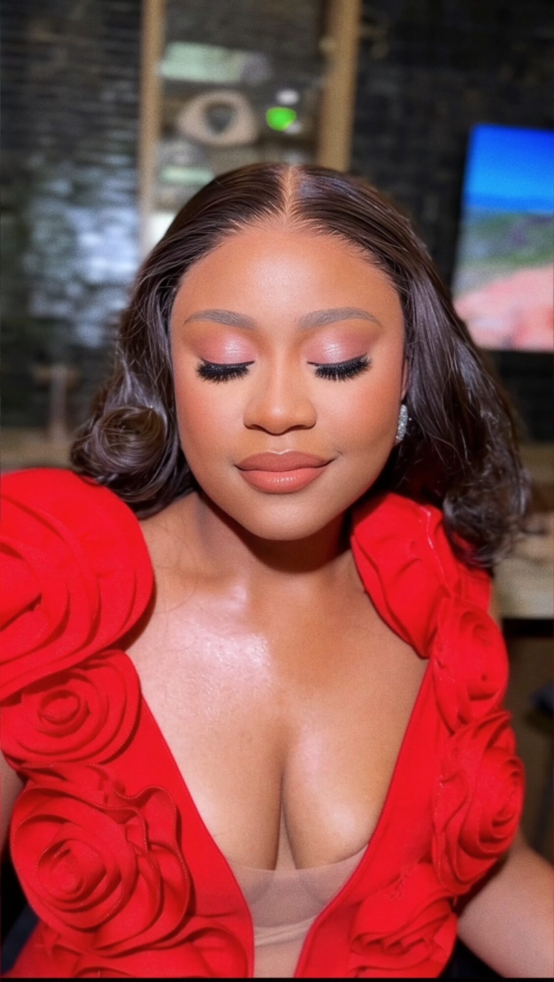 Woman with closed eyes and makeup, wearing a red dress with ruffled sleeves, in an indoor setting.