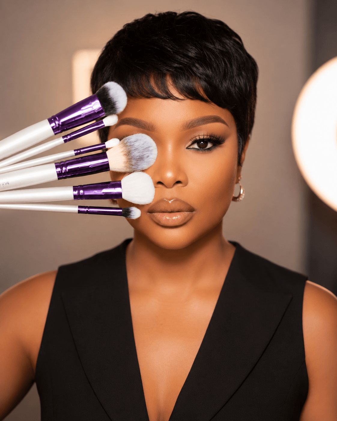 Woman with short black hair and makeup, surrounded by makeup brushes near her face, in a well-lit room.