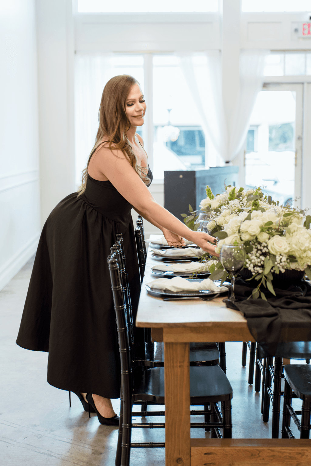 Woman in black dress arranging table settings with a large floral centerpiece in a bright room with large windows.