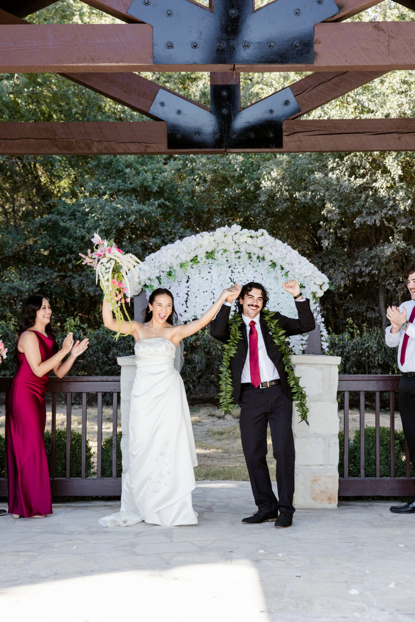 Bride and groom holding hands under an arch, celebrating with two women on a decorated outdoor stage.
