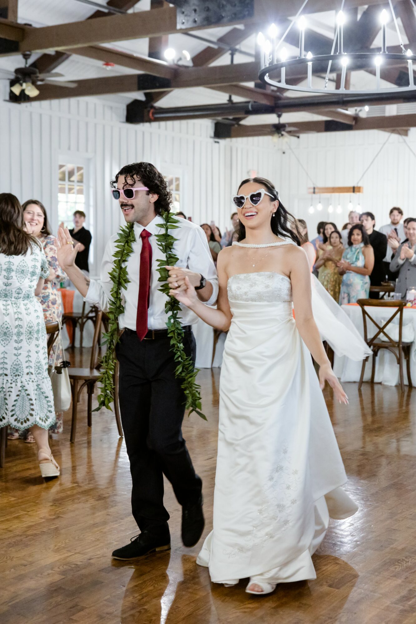 Bride and groom walking together in a wedding reception hall, surrounded by guests, wooden ceiling beams, and bright lighting.