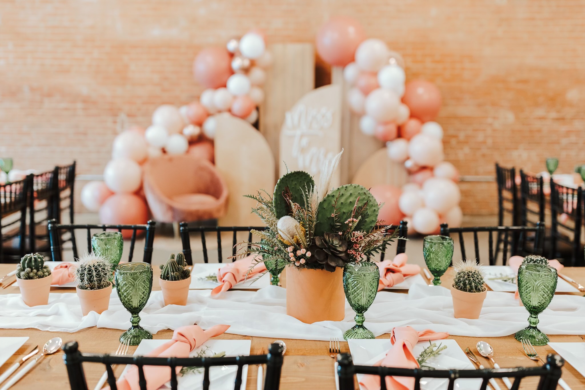 Decorated table with pink and green accents, balloons, and chairs for celebration, against a brick wall background.