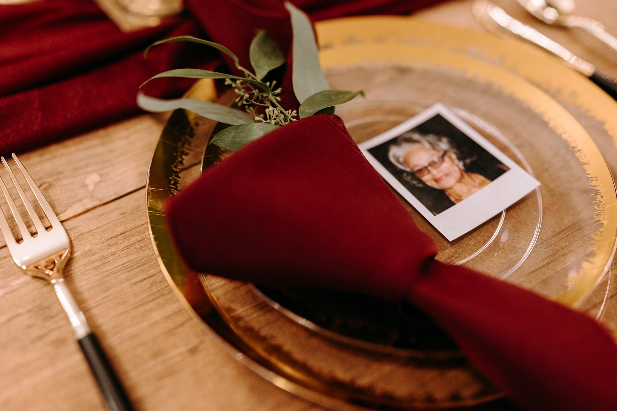 Table setting with a gold plate, burgundy napkin, a photo, and greenery, on a wooden table.