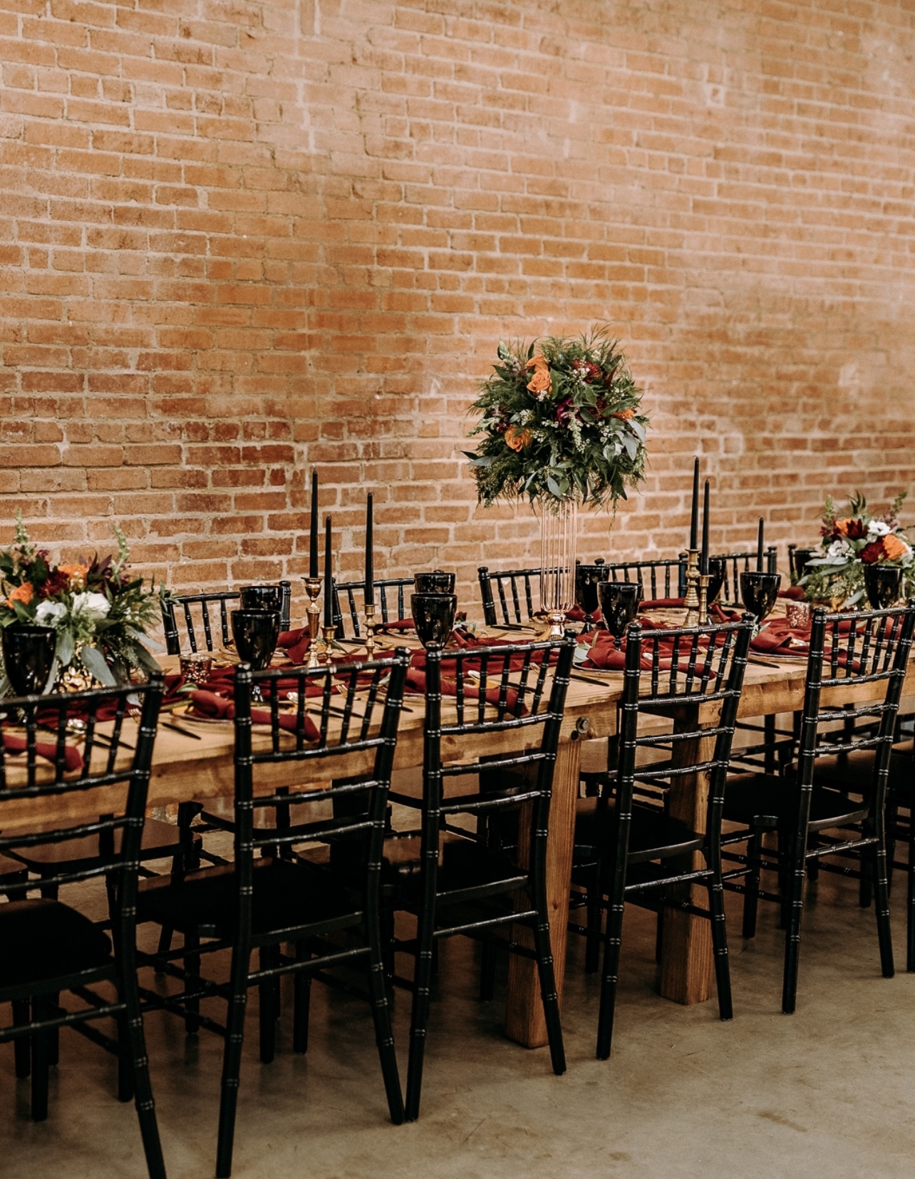 Long wooden table set with black chairs, floral centerpieces, and black candlesticks against a brick wall.