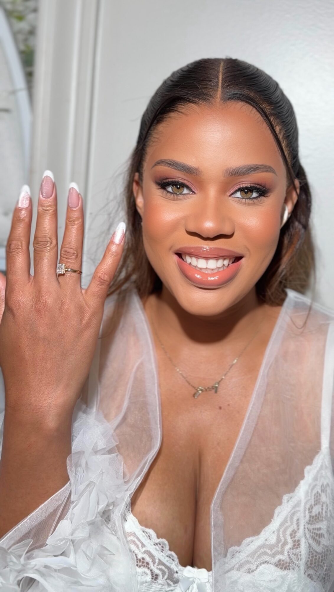 Woman smiling, showing a ring on her finger, wearing a white lace dress, with long dark hair and makeup.