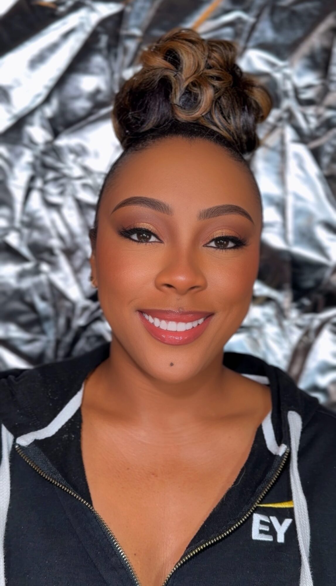 Smiling woman with styled hair and makeup, lying on shiny silver fabric, wearing a black zip-up top.