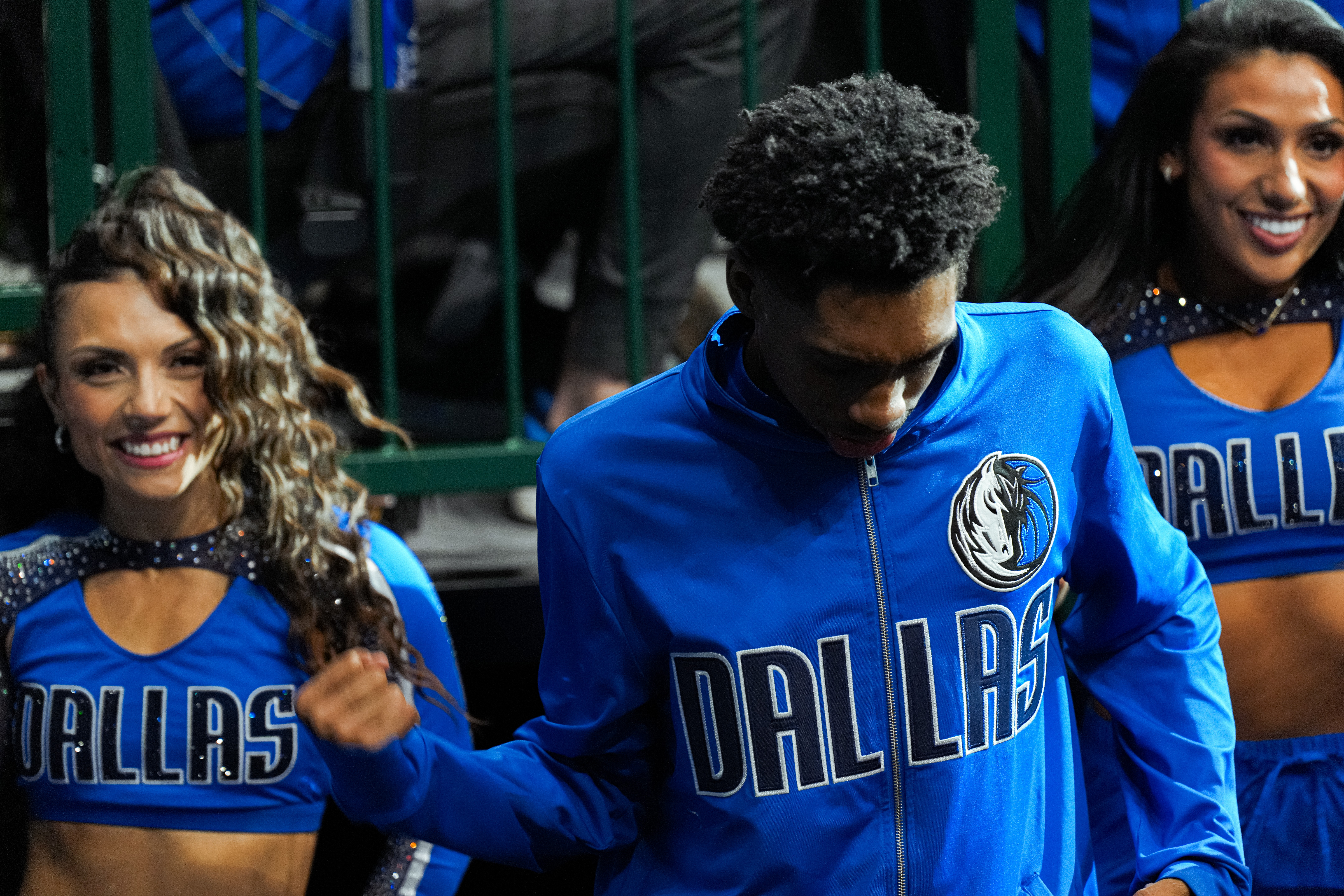 Three cheerleaders in blue Dallas uniforms smiling, with one adjusting her hair, behind green railings.