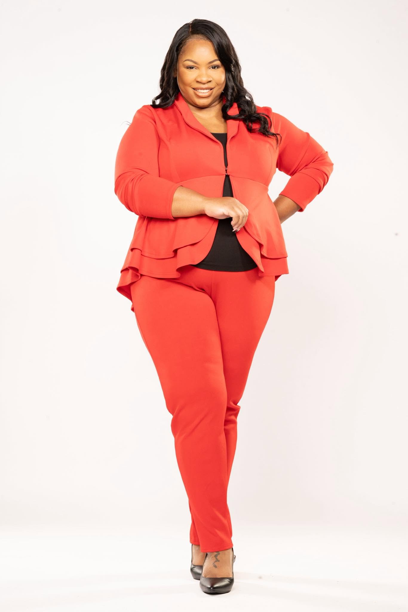 Woman in red suit with black top, smiling, standing with one hand on her hip, against a white background.