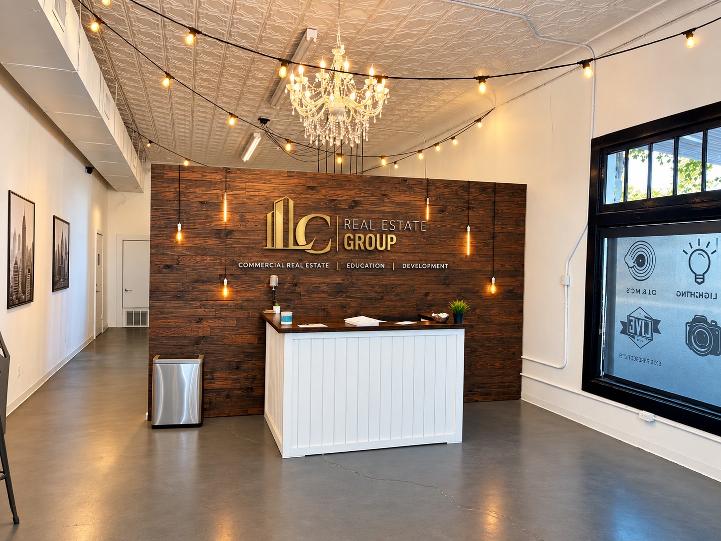 Reception area with a wooden wall, chandelier, string lights, and a white counter, in a modern office space.