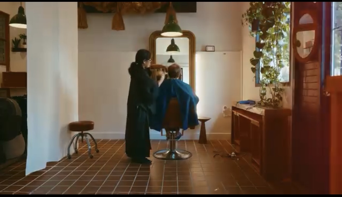 Two people in a room with a mirror, one sitting in a chair and the other standing nearby, with plants and furniture visible.