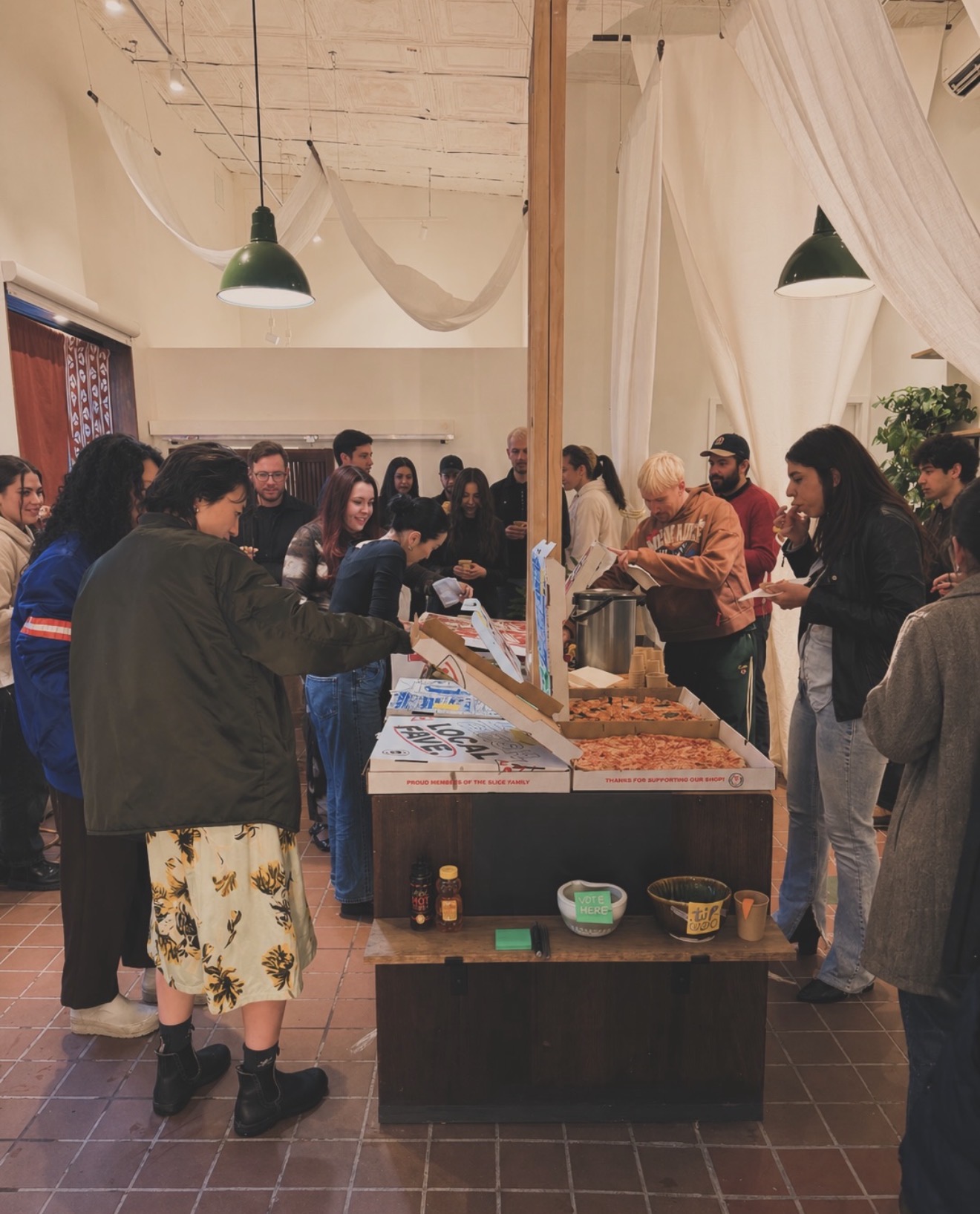 People gather around a food table in a spacious room with hanging lights and draped fabric, serving and socializing.