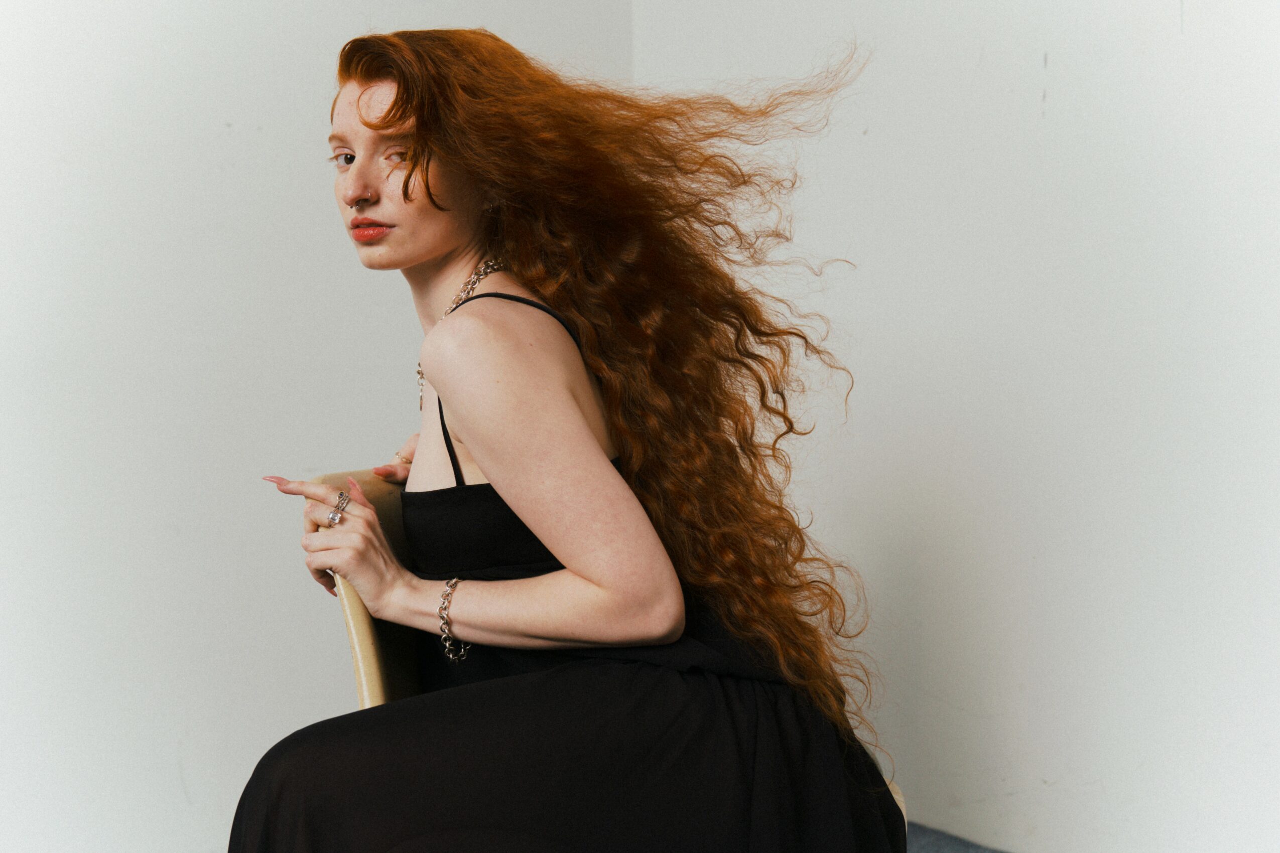 Woman with long, curly red hair wearing a black dress, standing sideways against a plain background.