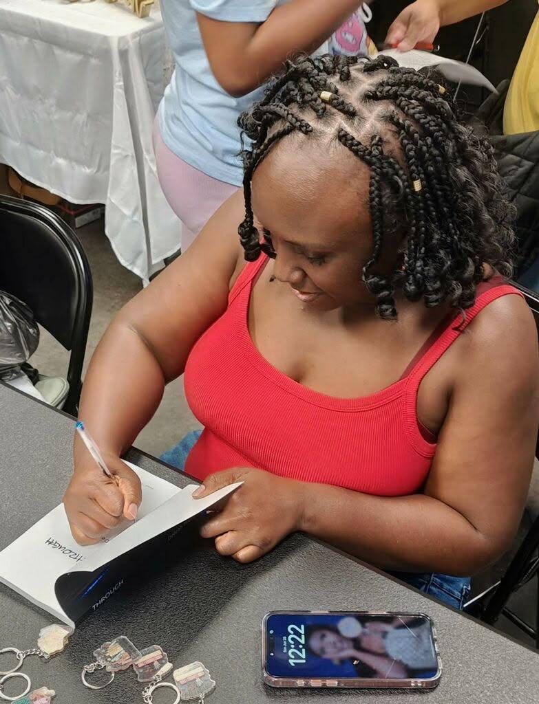 Woman with braided hair in a red top writing in a book at a table, with a smartphone nearby, in a social setting.
